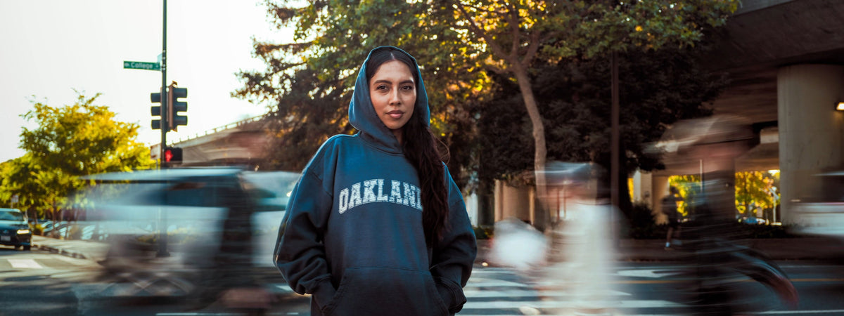 Person with curly hair wearing a black Oaklandish hoodie, standing outdoors with trees in the background. The sun sets in the distance, creating a warm glow in the sky. The person looks directly at the camera.