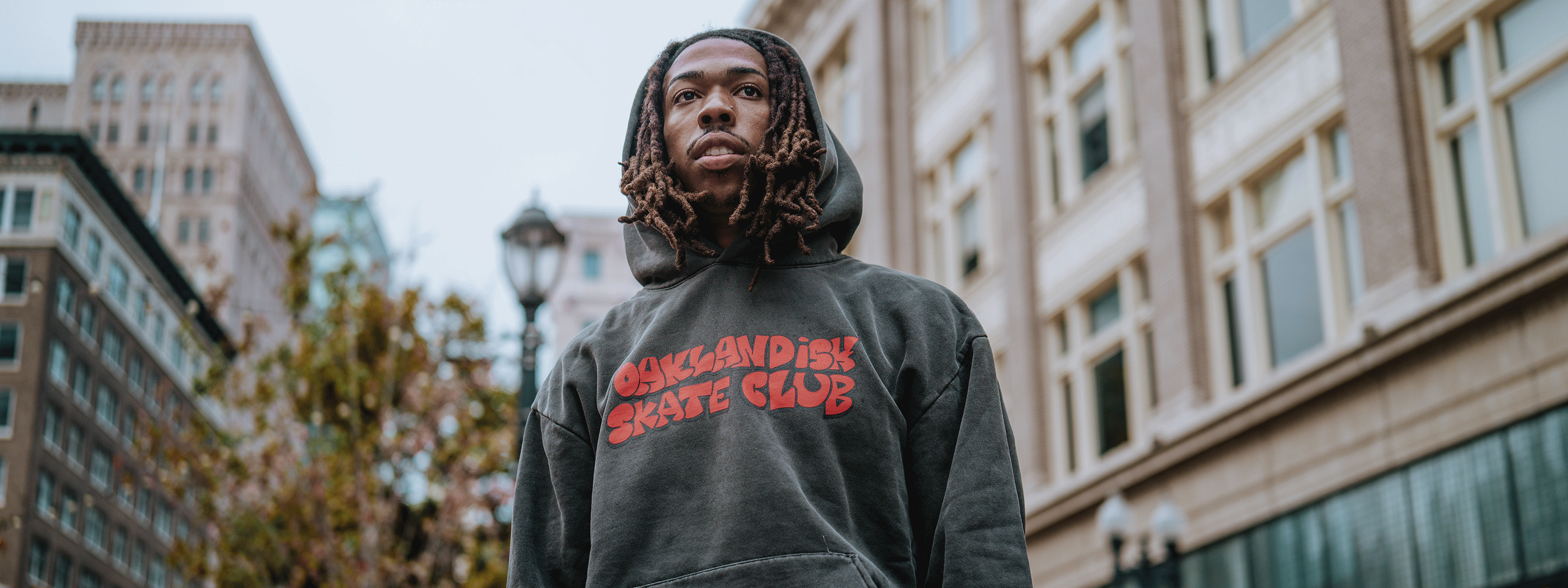 A young man with long dreadlocks wears a dark hoodie reading "Oaklandish Skate Club" in red letters, standing outdoors in a city with tall buildings and trees in the background.