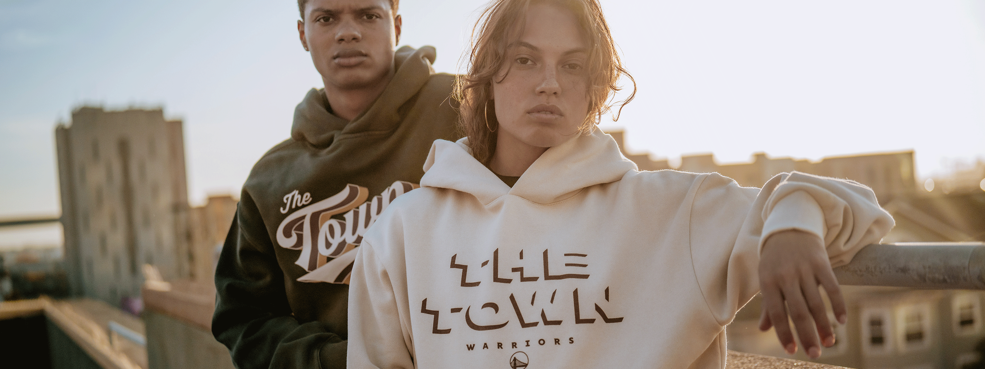 Two people stand on a rooftop at sunset, wearing hoodies—one black and one white—both with "The Town" and "Warriors" printed on the front in bold, stylized letters.