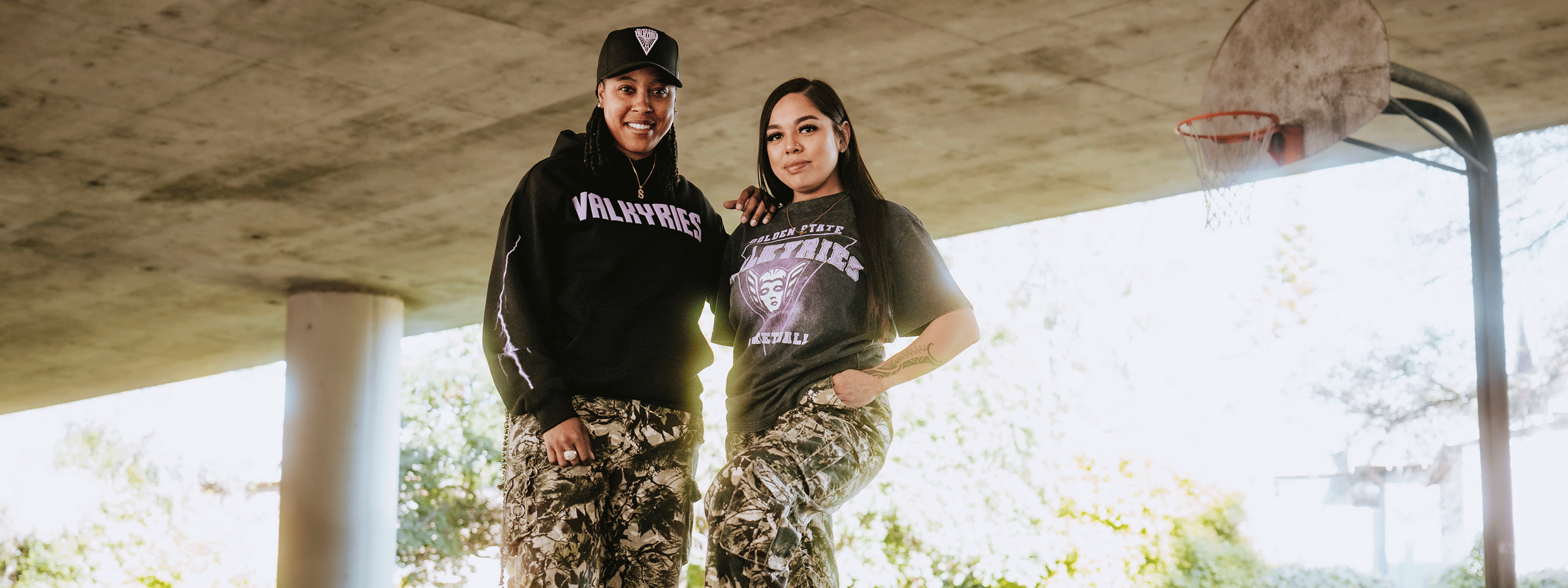 Two women in camo pants and “Valkyries” shirts stand confidently under a concrete overpass near a worn basketball hoop, smiling at the camera in natural daylight.