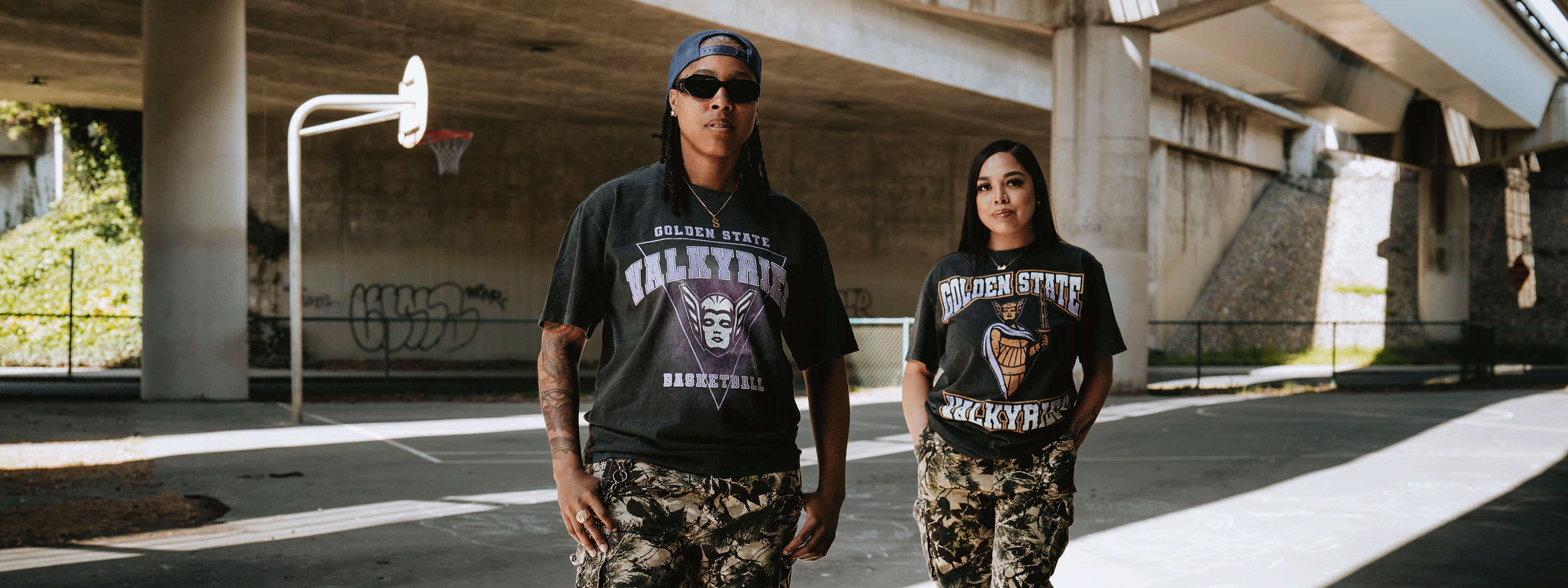 Two people in camo pants and graphic t-shirts stand on a sunlit, empty outdoor basketball court under a bridge; one wears sunglasses and a cap, both look confidently at the camera.