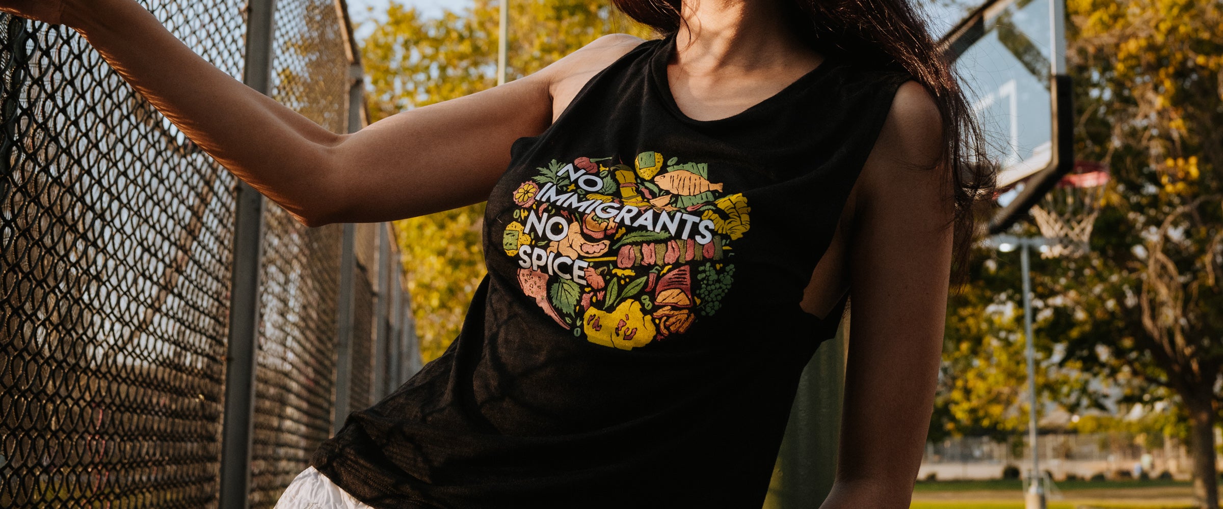 Cropped image of model wearing tank top with colorfull bbq plate of spice design, posing outside next to fence.