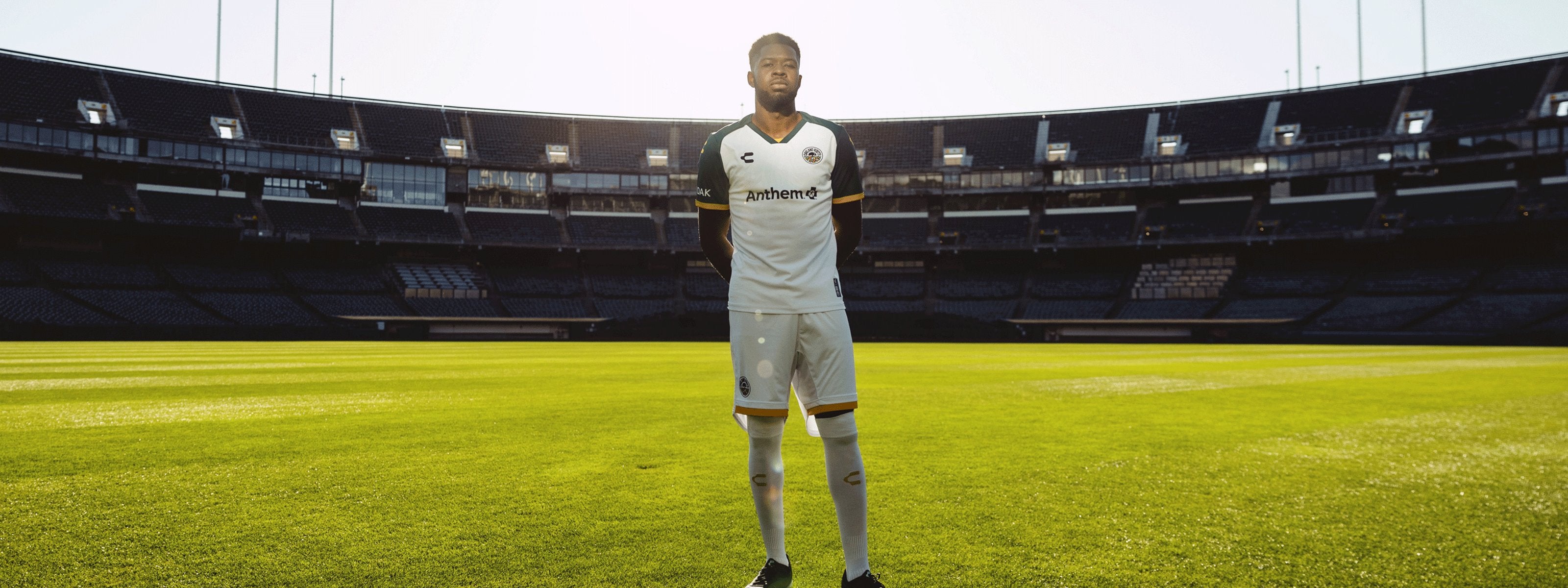 Soccer player in a white and black jersey stands on a sunlit soccer field in an empty stadium, arms behind his back. The sunlight creates a halo effect behind him, highlighting the green grass.