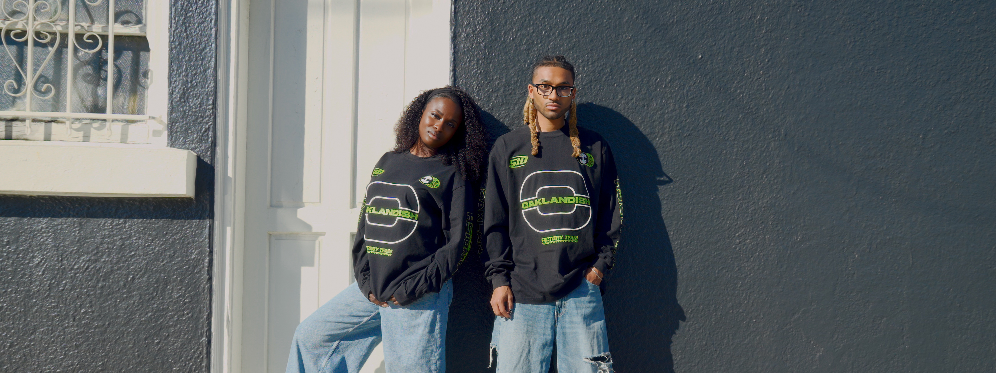 A woman and man stand by a lake, wearing matching "Make America Native Again" sweatshirts. The background features a grassy shore and distant buildings under a cloudy sky.