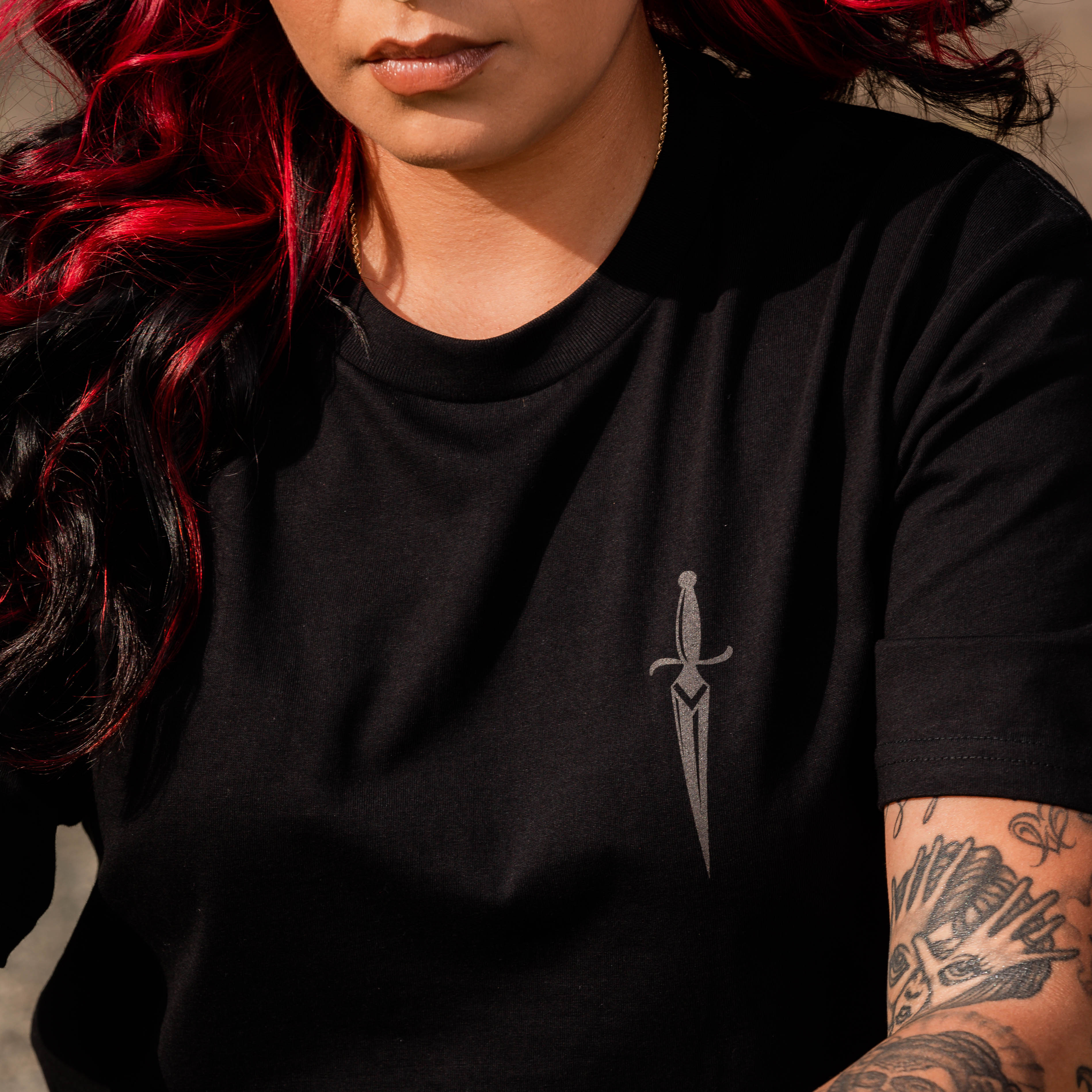 A person with long, dark hair featuring red highlights is wearing the Blossom Tee by Oaklandish—a men's cotton t-shirt adorned with a small sword graphic on the left side of the chest. Their left arm is decorated with various tattoos, including floral motifs and other intricate patterns. The person is sitting outdoors.