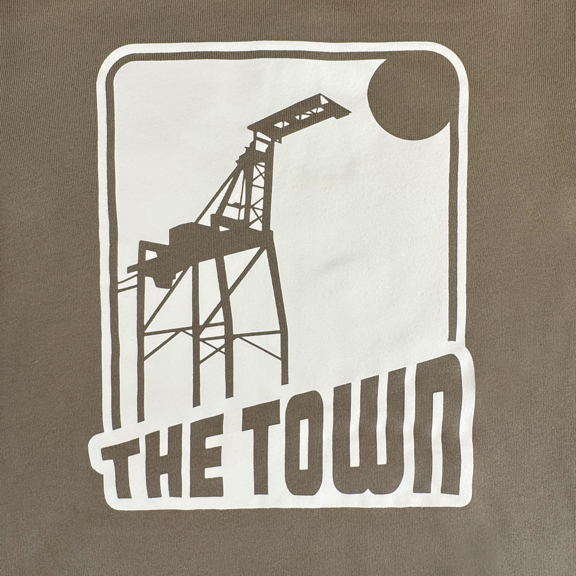 A black and white graphic of a large crane with a sun or moon in the corner sits above bold "THE TOWN" text. This 70’s Trading Card Zip Hoodie by GSW features an Oaklandish design inside a rectangular frame on a brownish background.