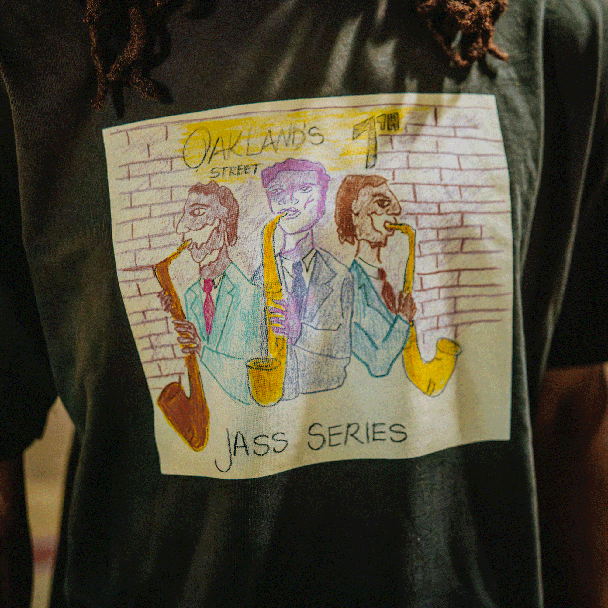 The Oaklandish 7th St. Players Tee is a black, 100% cotton classic fit t-shirt featuring the "JASS SERIES" hand-drawn art of three men in suits playing saxophones, with vibrant yellow, blue, and red accents against a brick wall.