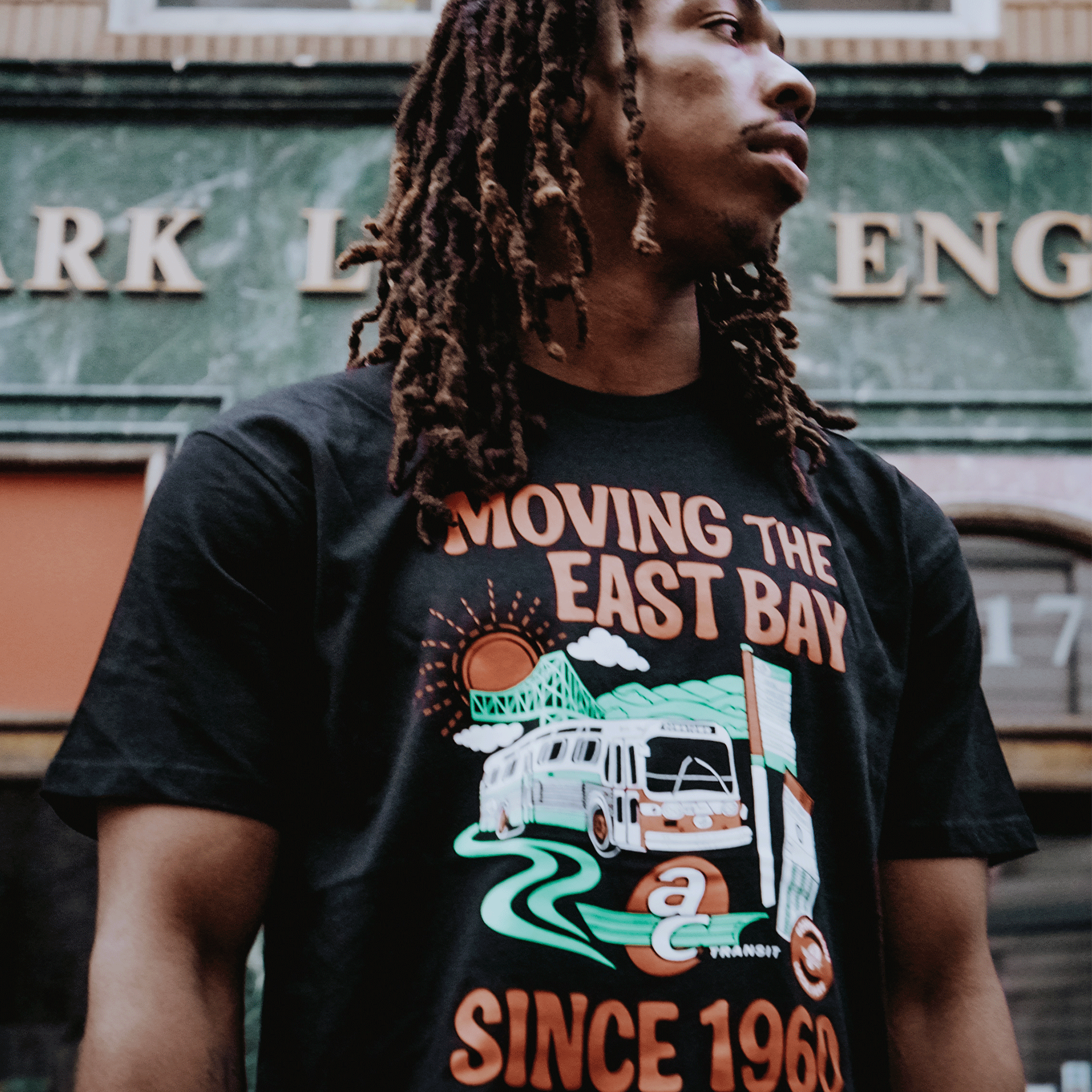 A young man with long, brown dreadlocks looks away from the camera, wearing the Oaklandish AC Transit Moves The East Bay Tee. A green marble building with gold lettering is visible in the background.