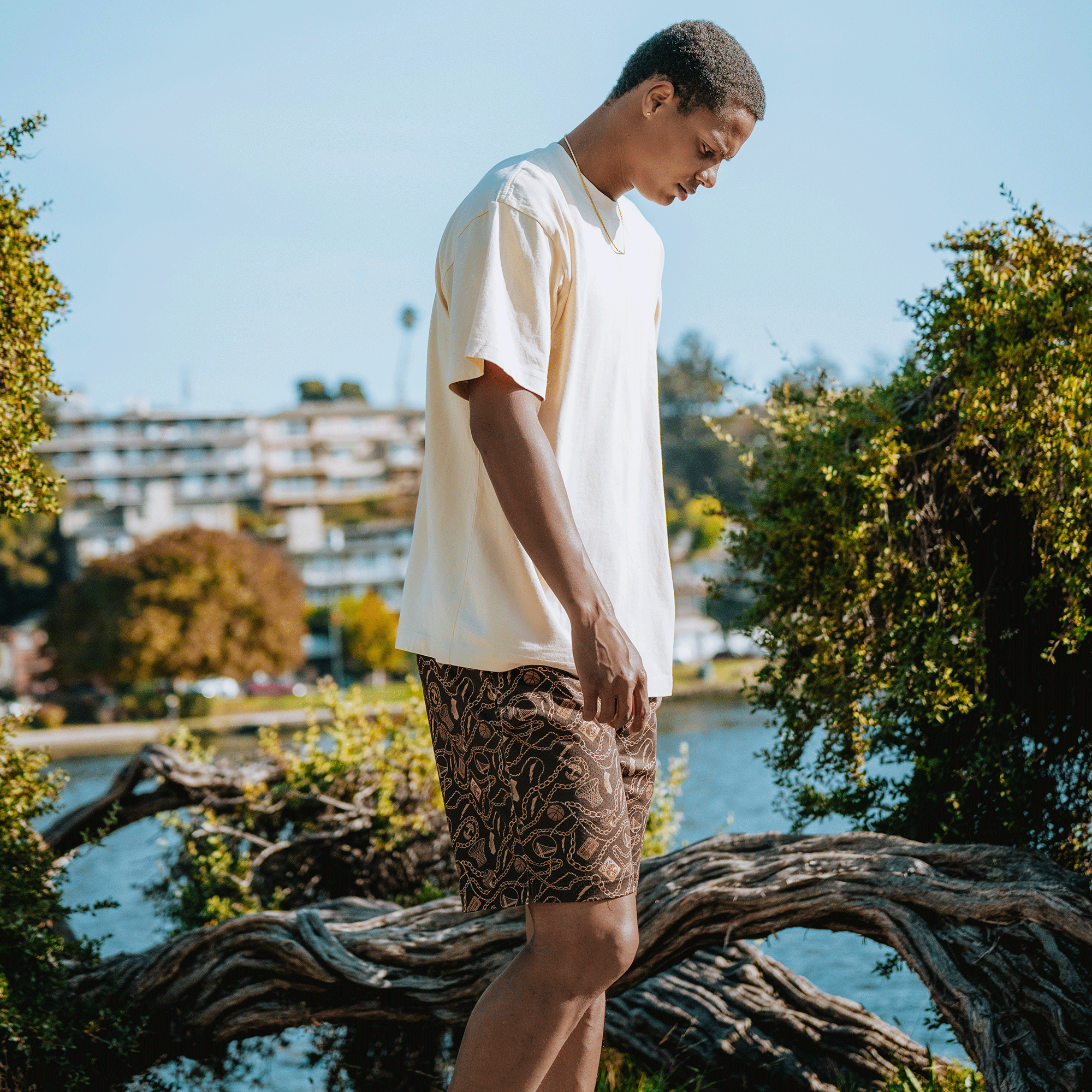 A man in a loose white T-shirt and GSW All Gold Forever Shorts stands outdoors by a twisted tree branch, with water, greenery, and blurred buildings behind him under a clear blue sky, looking down thoughtfully.
