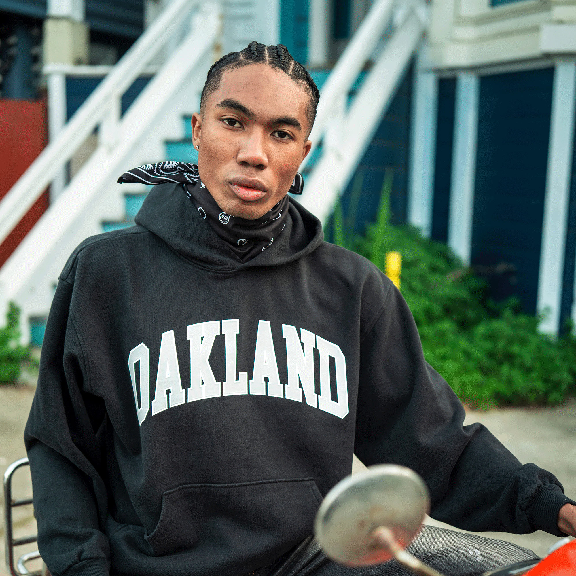 A young person with braids, wearing an Oaklandish Alma Mater Hoodie 2.0 and a black bandana, sits outdoors near white steps and blue buildings holding a fencing sword, with green plants and a yellow pole in the background.