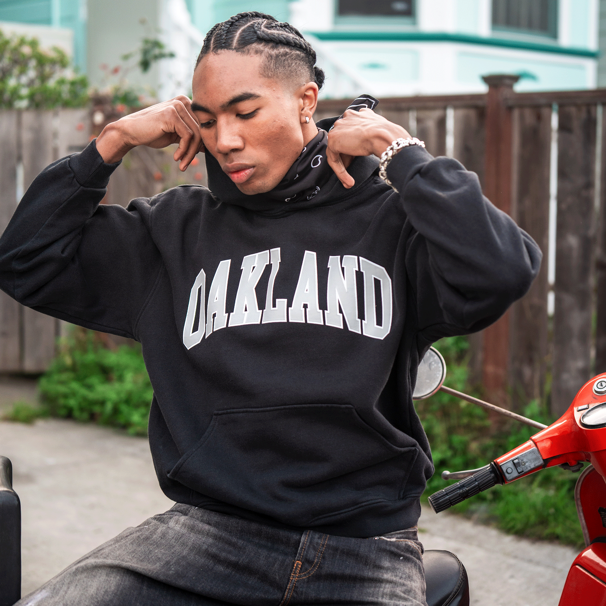 A young man with braided hair sits on a red scooter, wearing the oversized Oaklandish Alma Mater Hoodie 2.0 and a silver bracelet. He lifts the hood, relaxed by a wooden fence with a pastel house behind him—a casual urban vibe.