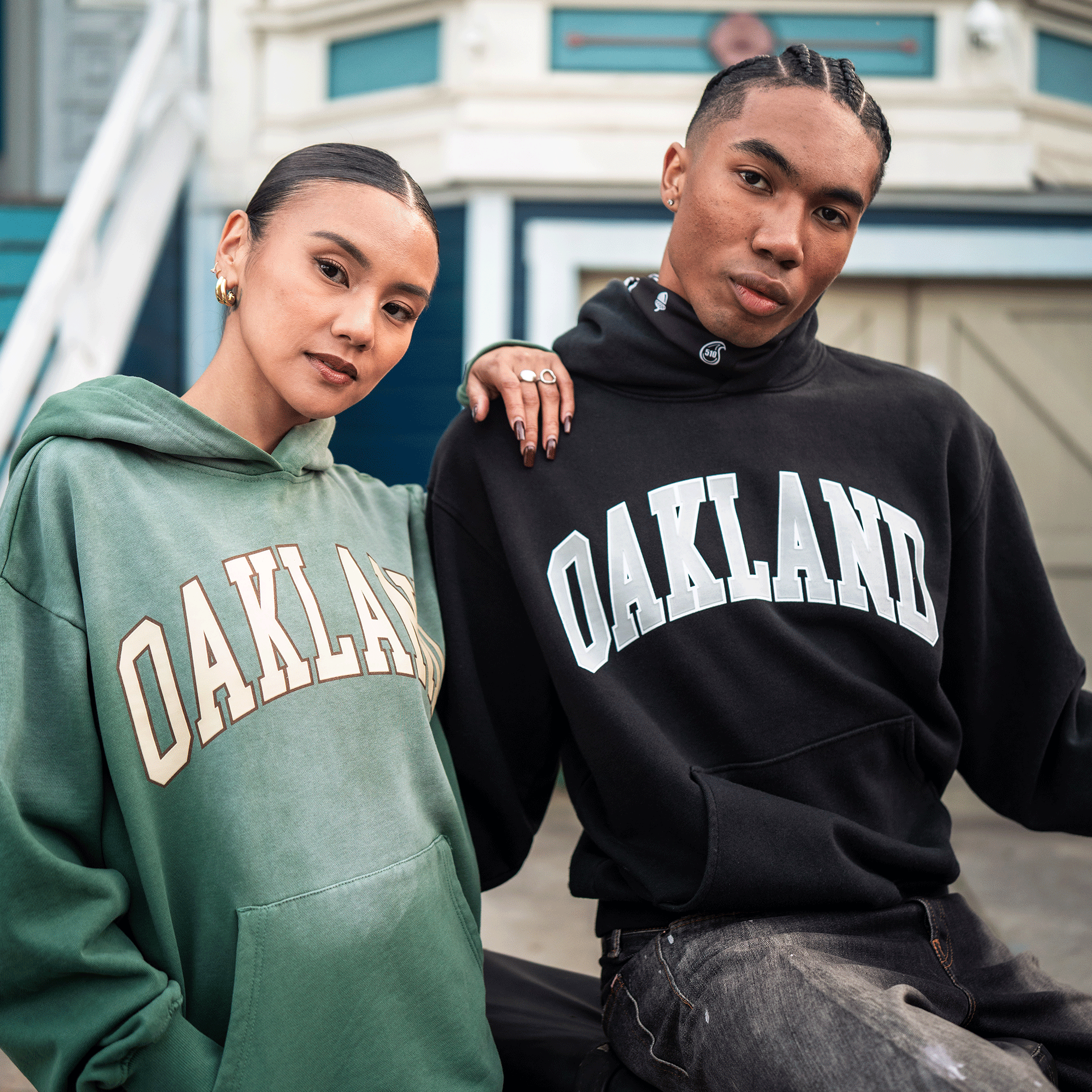 Two young adults sit closely, wearing Oaklandish Alma Mater Hoodie 2.0 styles in green and black. The woman pairs hers with gold hoops; the man sports rings. Both exude confidence outdoors in these 100% cotton hoodies.