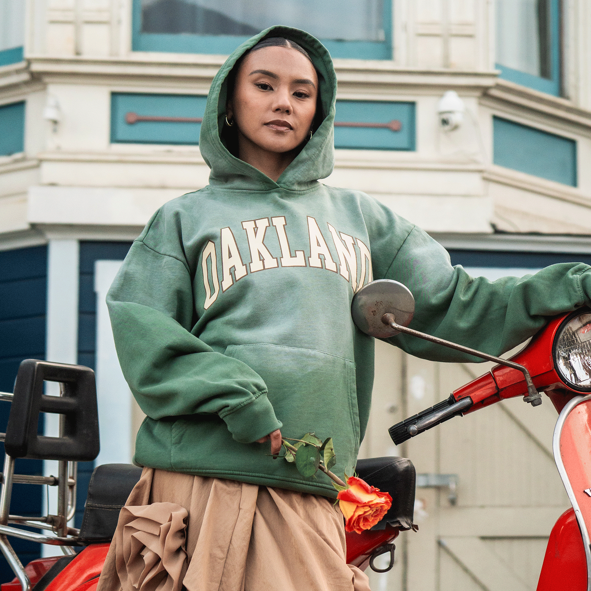 A woman in an oversized green Oaklandish Alma Mater Hoodie 2.0 and tan skirt poses confidently by a red scooter, one hand on the handlebars, in front of a blue and white building, looking directly at the camera.
