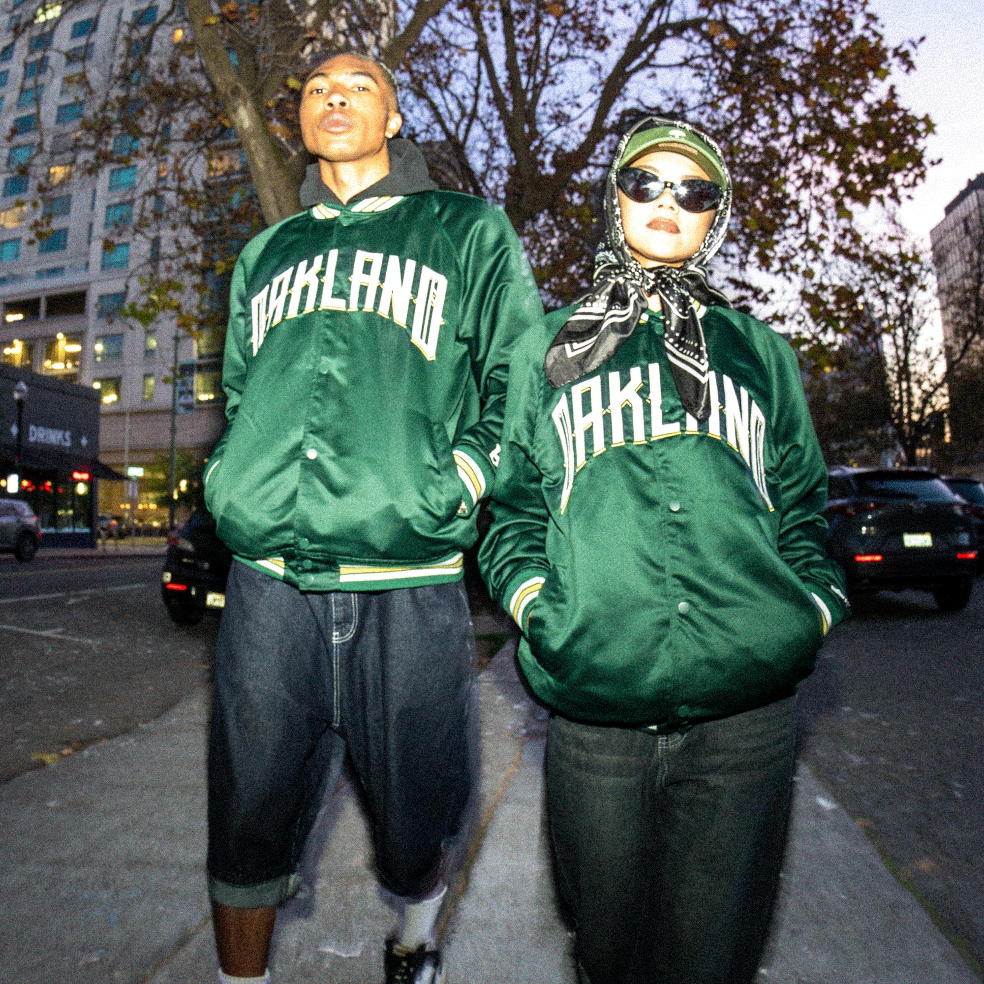 Two people pose on a city sidewalk in matching Oakland Ballers Satin Jackets by Oakland Ballers. One wears shorts and a cap, the other sunglasses, scarf, and pants. Autumn leaves and buildings set a lively urban scene with a stylish, slightly blurred vibe.