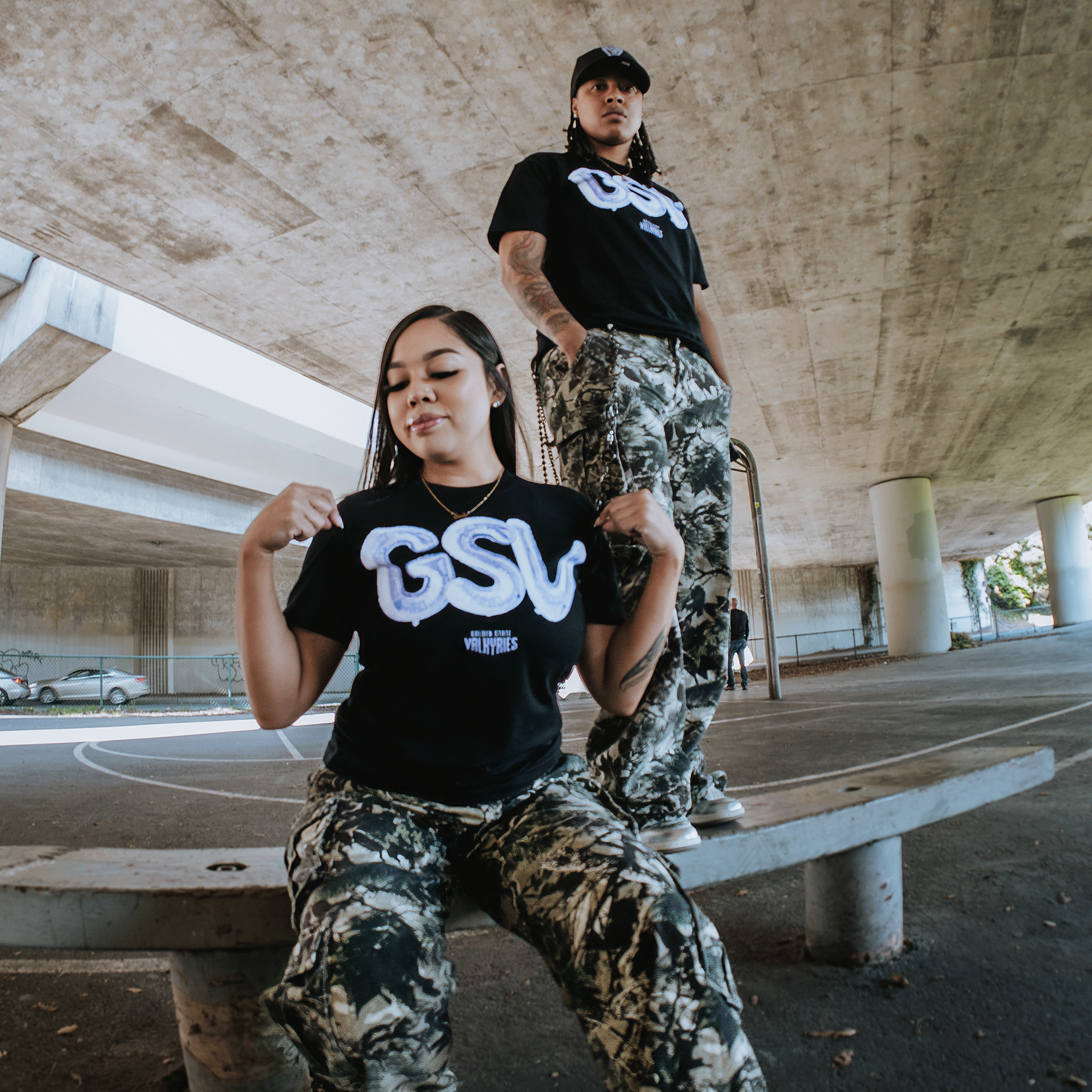 Two people in matching black GSV Golden State Valkyries Balloons Tees and camo pants pose under a concrete overpass—one sits tugging her shirt on a curved bench, while the other stands on an urban basketball court, channeling Valkyries energy.
