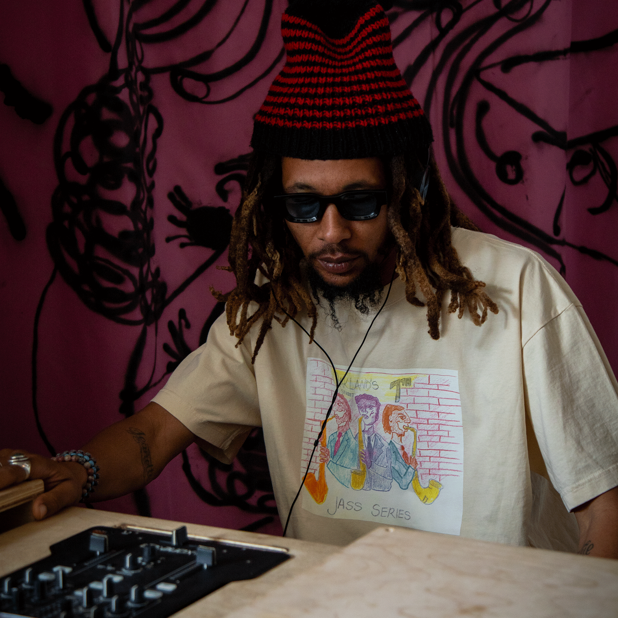 A man with long dreadlocks and a red-and-black knit hat sits at a DJ controller, wearing dark sunglasses and the Oaklandish 7th St. Players Tee—a classic fit, 100% cotton shirt with an illustrated design—against a vibrant pink wall with black abstract patterns.