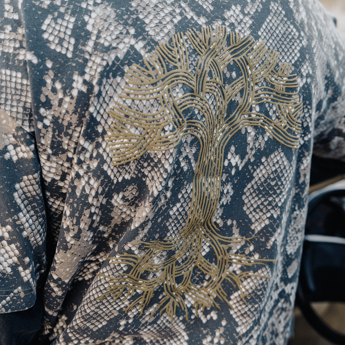 The Oaklandish Classic Logo LS Tee is a classic fit long-sleeve shirt in a gray and black snakeskin print, featuring a large, stylized olive green oak tree graphic on the back drawn with fine, continuous lines depicting branches and roots.