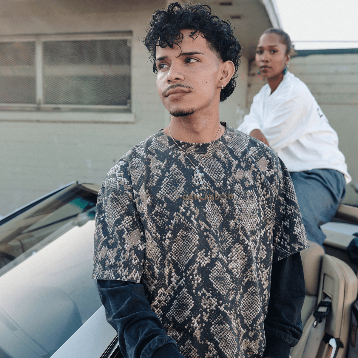 A person with curly hair and a mustache wears a dark snake-print shirt layered over an Oaklandish Classic Logo LS Tee, standing beside a car and another person in a white top in front of a neutral-colored building.