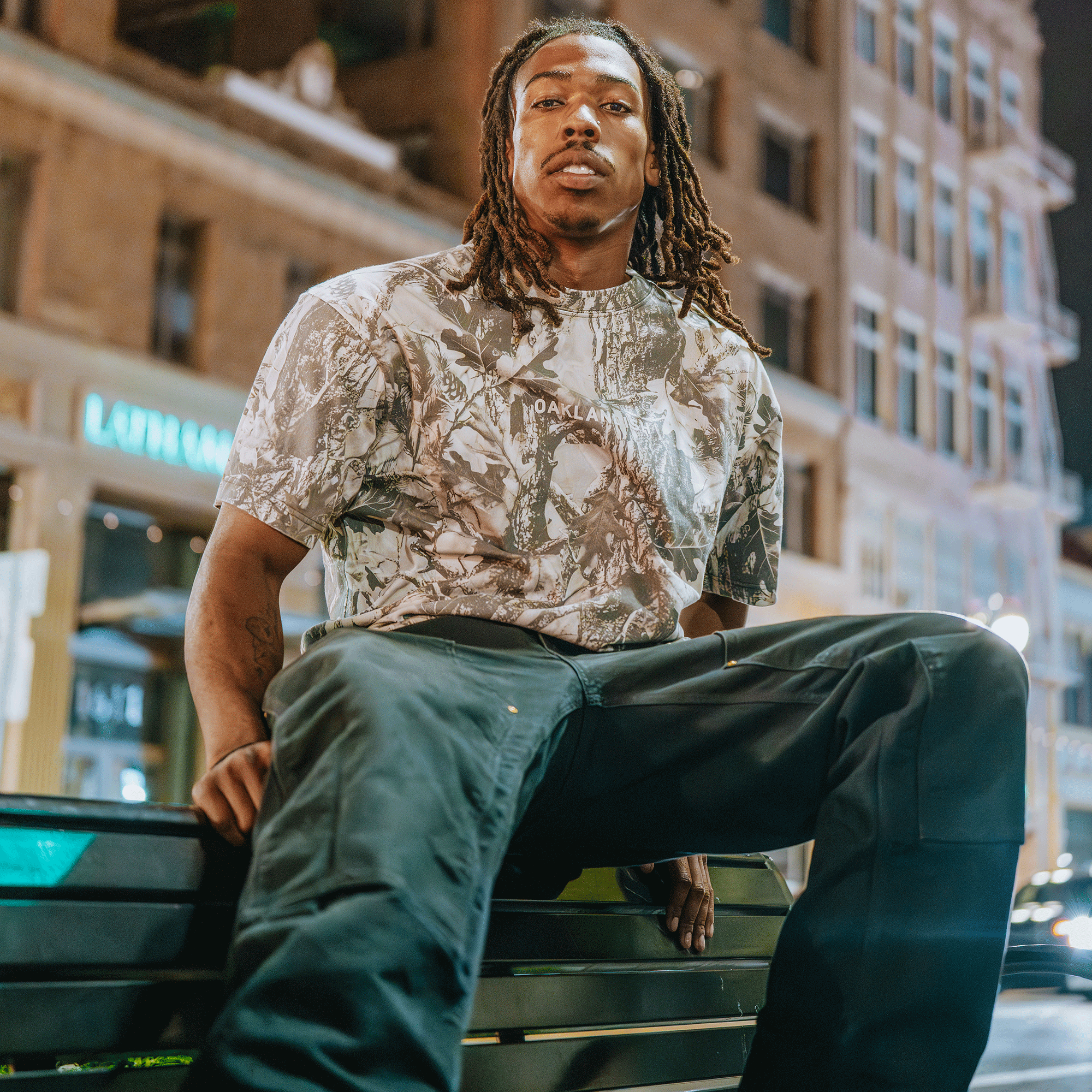 A man with long dreadlocks sits on a city bench at night, illuminated by bright lights. He confidently looks at the camera, wearing the Oaklandish Classic Logo Tee by Oaklandish.