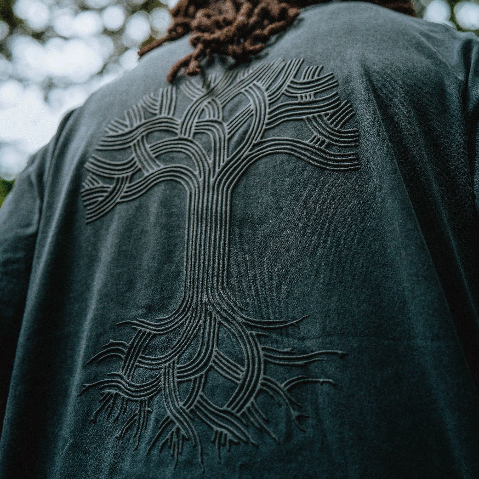A close-up of the Oaklandish Classic Logo Tee by Oaklandish, a men's cotton t-shirt in a classic fit with an intricate, raised tree design, worn by a person with dreadlocks standing outdoors against blurred greenery.