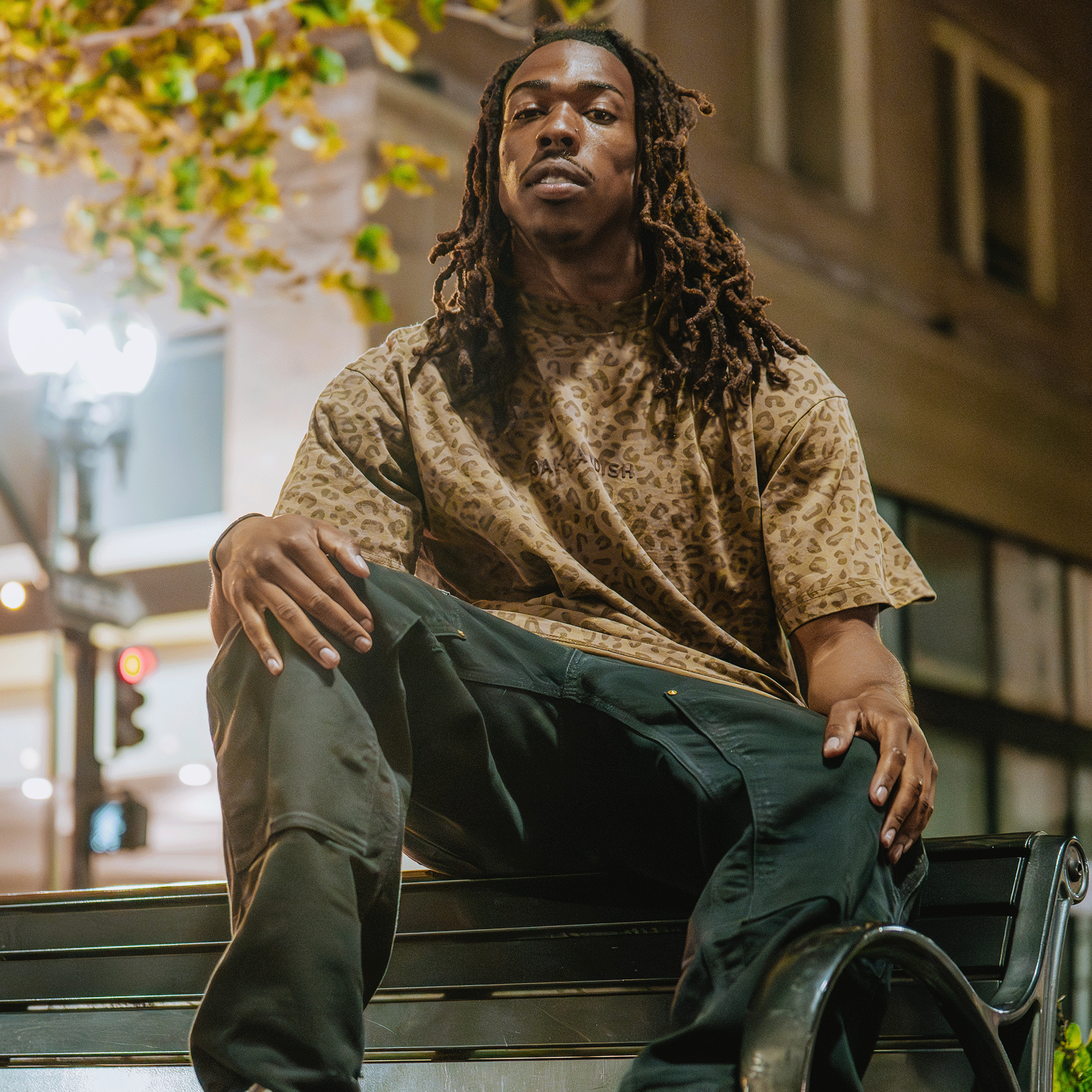 A young man with long dreadlocks sits confidently on a bench at night, wearing the Oaklandish Classic Logo Tee by Oaklandish and dark pants. City streetlights, a building, and green leaves blur softly in the urban background.