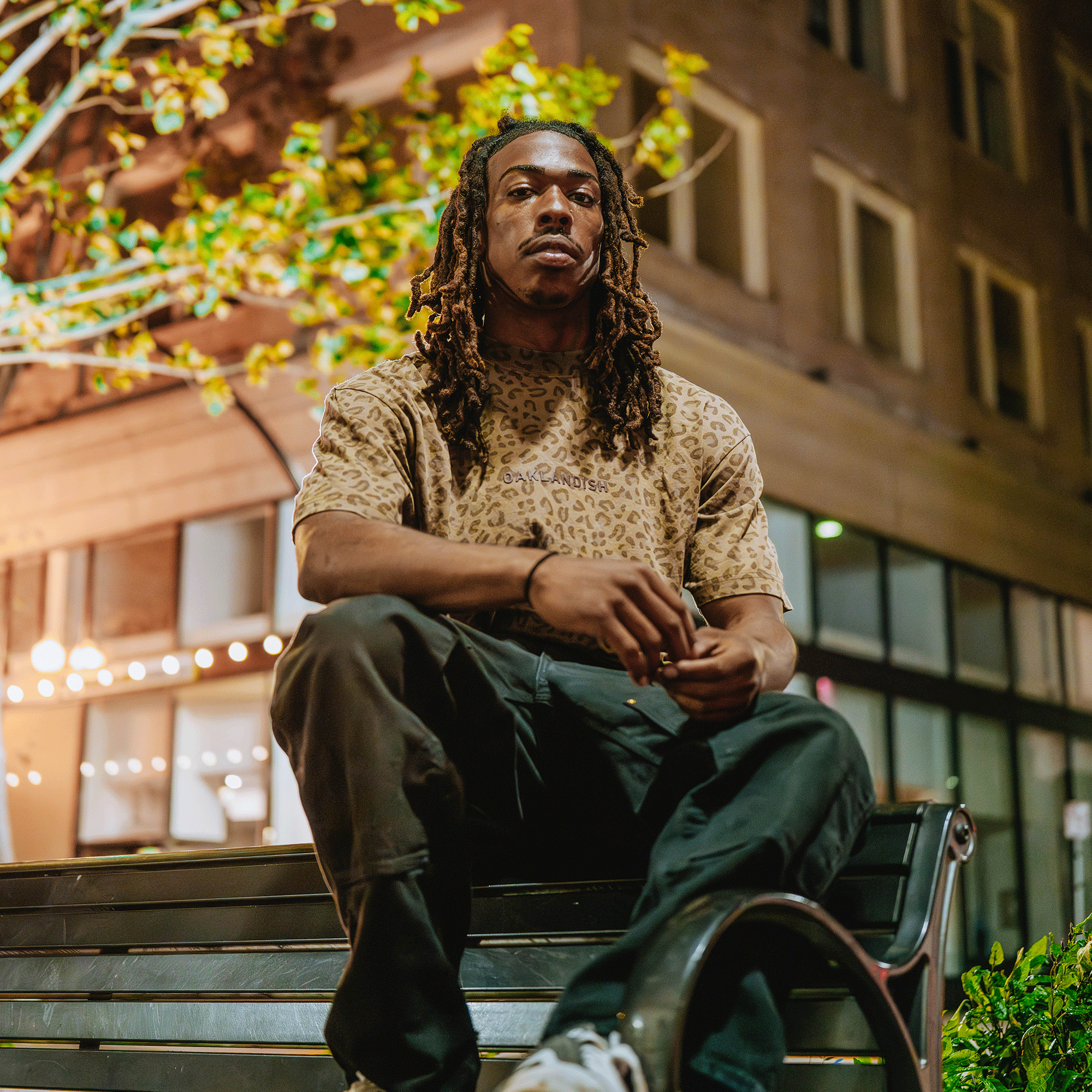 A man with long dreadlocks sits on a bench at night, wearing dark pants and an Oaklandish Classic Logo Tee by Oaklandish. Illuminated windows, string lights, and leafy branches behind him create a relaxed urban vibe.