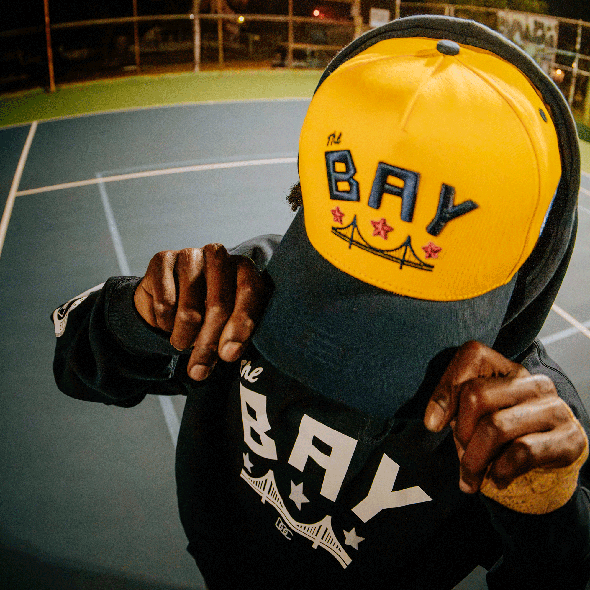 On a blue outdoor court at night, someone in a black hoodie adjusts the brim of their yellow Bay Bridge Snapback by DOC from Oaklandish, its bridge graphic reflecting Bay Area global culture, with their face mostly hidden under the hat.
