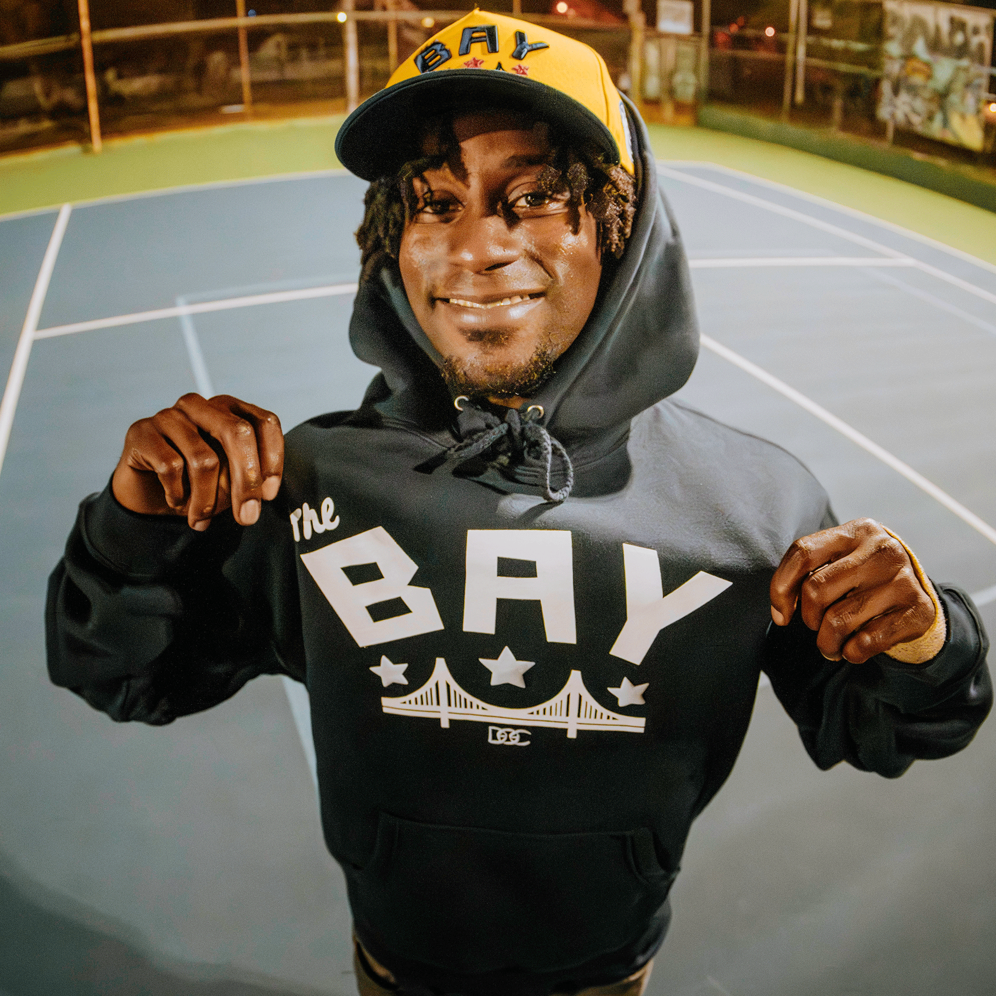 A smiling person stands on a tennis court at night, wearing the Bay Bridge Hoodie by DOC from Oaklandish—a navy hoodie and cap with “The BAY” and bridge graphics—showing off Bay Area pride under bright court lights.
