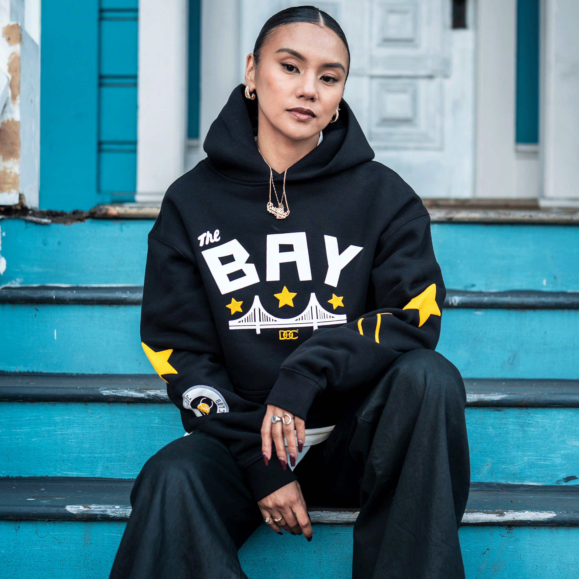 A woman with slicked-back hair sits on turquoise steps, confidently wearing the Bay Bridge Hoodie By DOC V2 from Oaklandish, paired with black pants and gold jewelry, set against a blue building and white door backdrop.