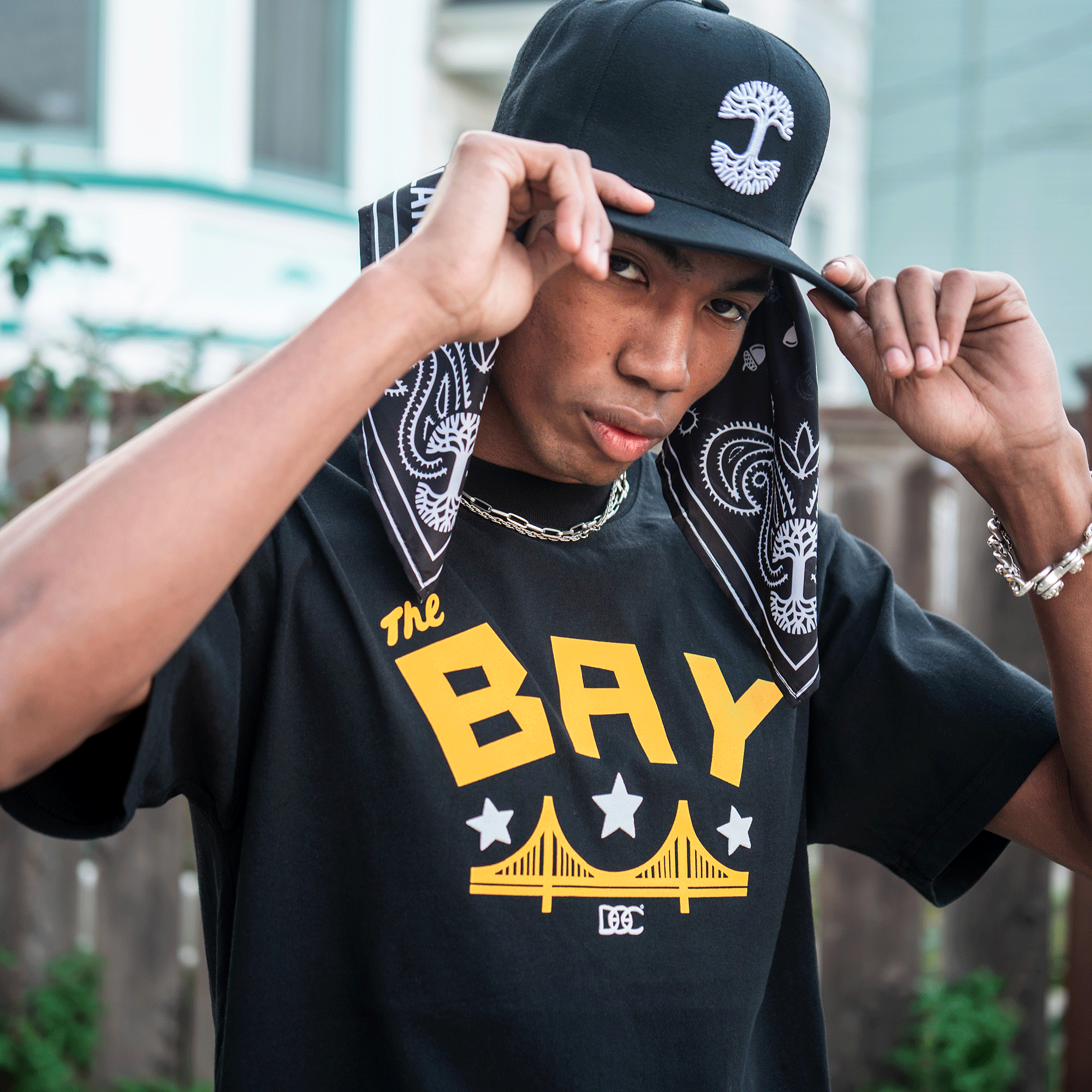 A young man in men's sizing wears a black cap, bandana, and chain. He lifts his hat, showing the Oaklandish Bay Bridge Tee By DOC V2 with "The BAY" above a yellow bridge graphic. A fence and house blur in the background.