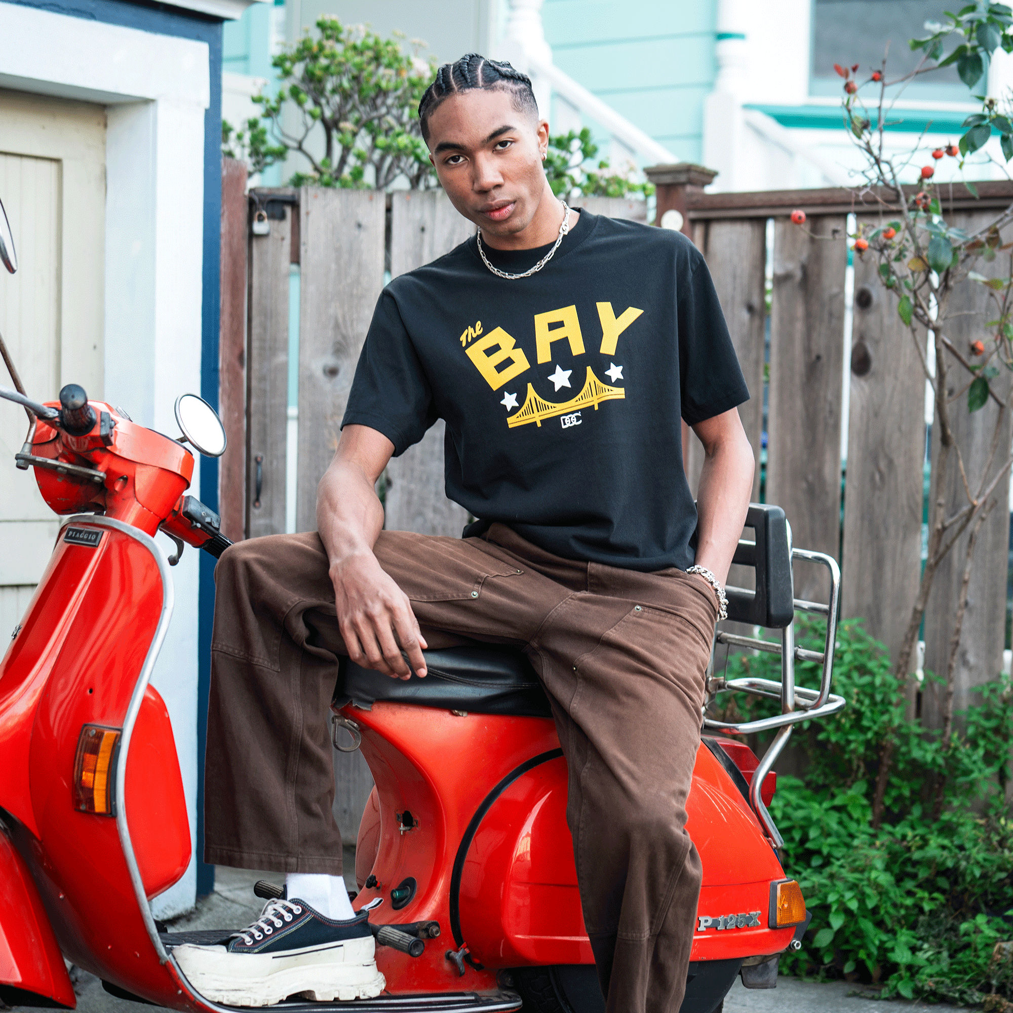 A young man with braided hair sits confidently on a red scooter, wearing the Oaklandish Bay Bridge Tee By DOC V2 in men's sizing, paired with brown pants and black-and-white sneakers. Behind him are a wooden fence, greenery, and a light blue house.
