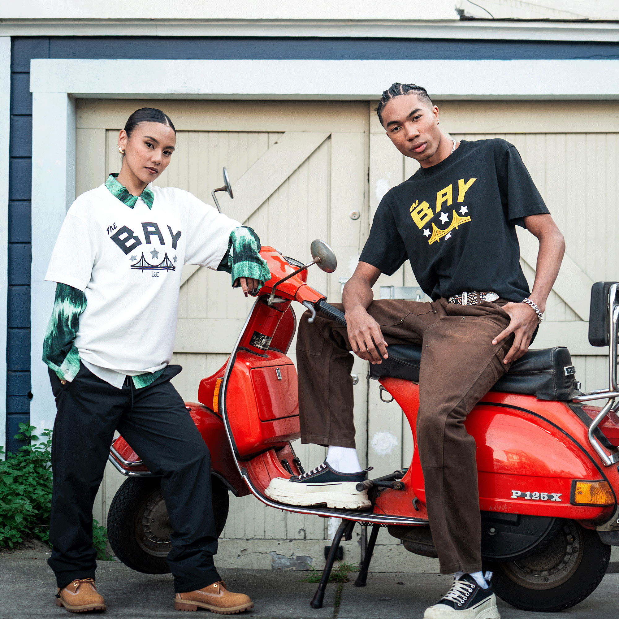 Two young adults pose by a red vintage scooter, each wearing the Bay Bridge Tee By DOC V2 from Oaklandish—one in white over green plaid, the other in black—highlighting modern men's heavyweight cotton style.