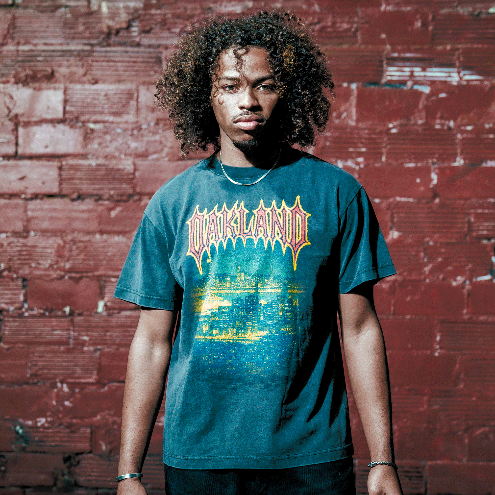 A young man with curly hair stands by a red brick wall, wearing the Oaklandish Ember Tee—a men's black t-shirt with bold yellow "OAKLAND" lettering. He gazes seriously at the camera, hands at his sides, with a chain around his neck.