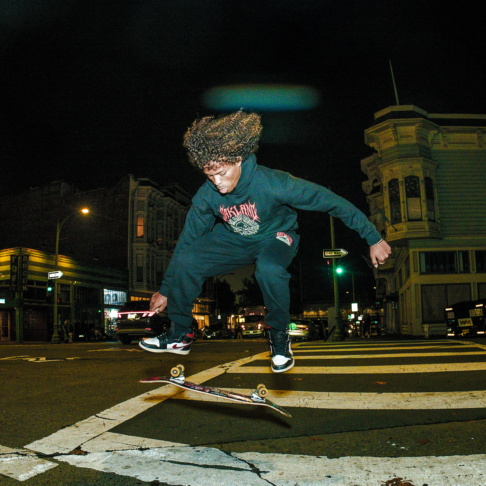 A young person with curly hair, wearing an Oakland Roots SC Forge Hoodie and black pants, performs a skateboard trick at night above a city crosswalk, with historic buildings and glowing streetlights in the background.