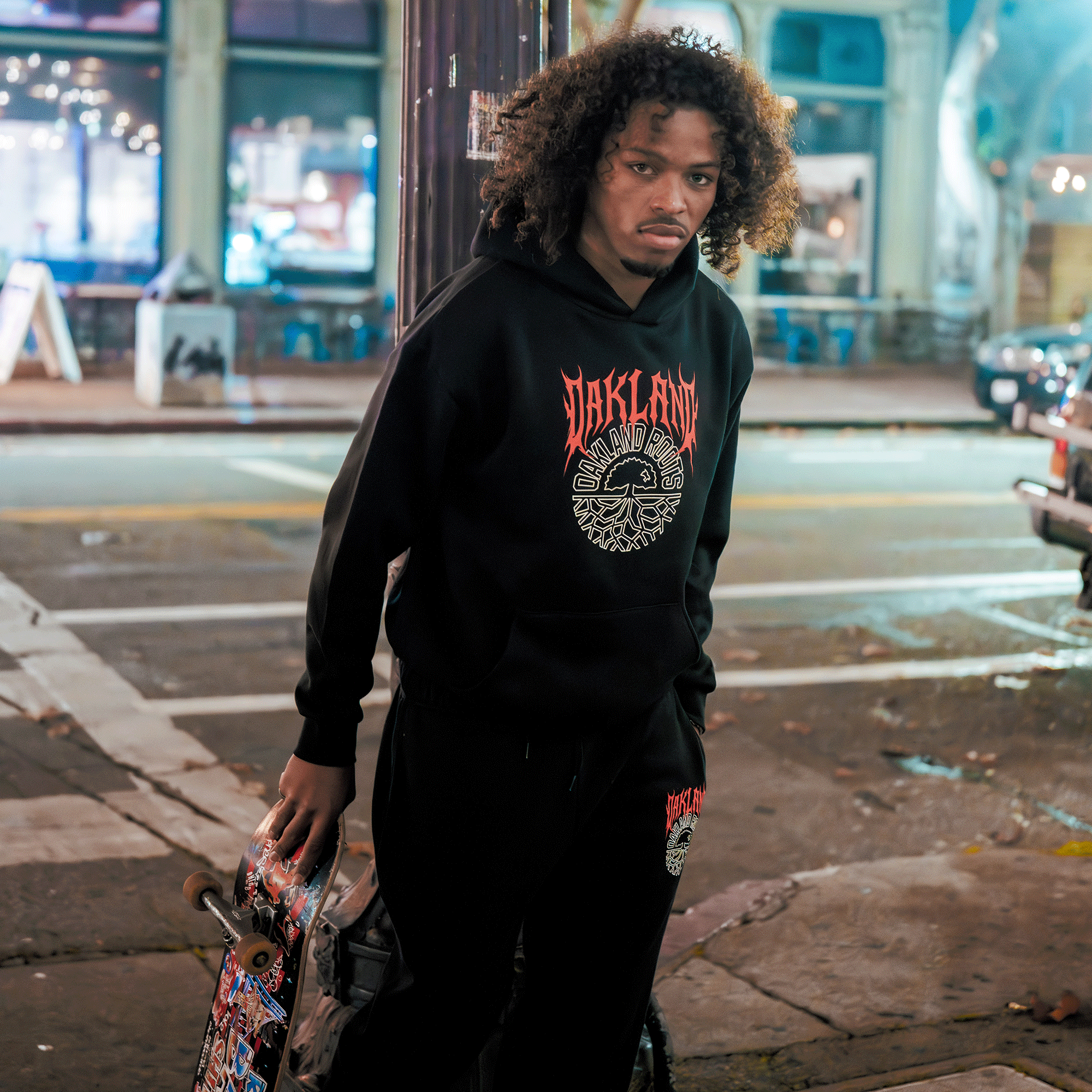 A young man with curly hair stands on a city sidewalk at night, holding a skateboard and wearing the Oakland Roots SC Forge Hoodie. Behind him are a street, parked cars, and brightly lit storefronts with large windows.