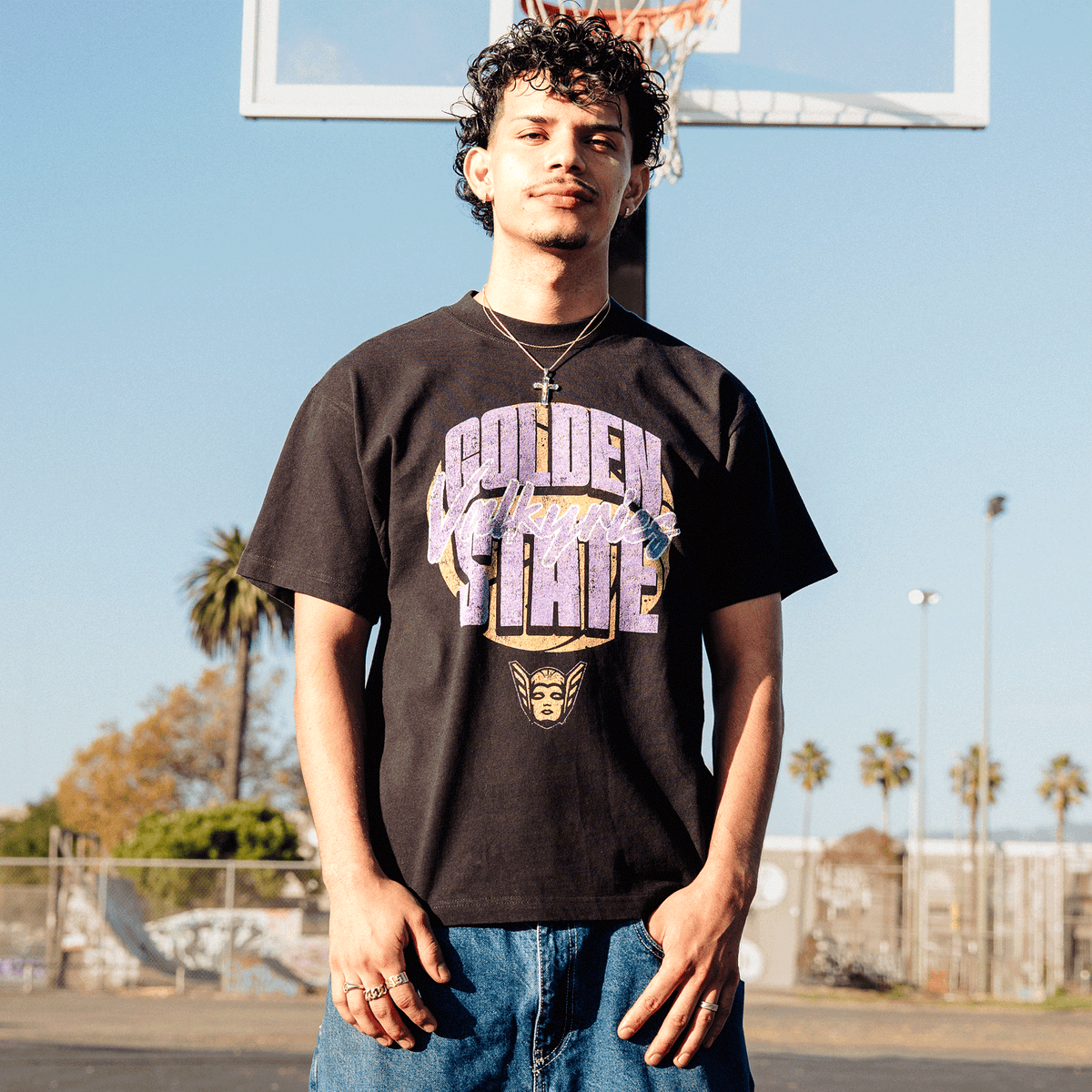 A young man with curly dark hair and a thin mustache wears a GSV Fade Away Tee and blue jeans on an outdoor basketball court, with the black shirt featuring large, weathered purple and yellow “Golden State” text above a small winged helmet icon.