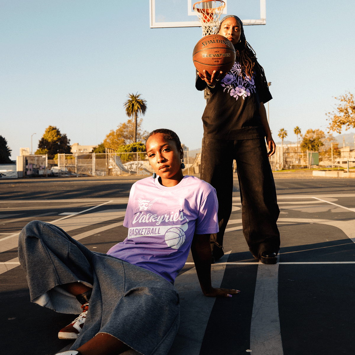 Two young Black women pose on an outdoor basketball court; one sits in a lavender classic fit t-shirt and grey wide-leg pants, while the other stands behind her wearing a black GSV Vintage Tee and baggy black pants, holding a basketball toward the camera.