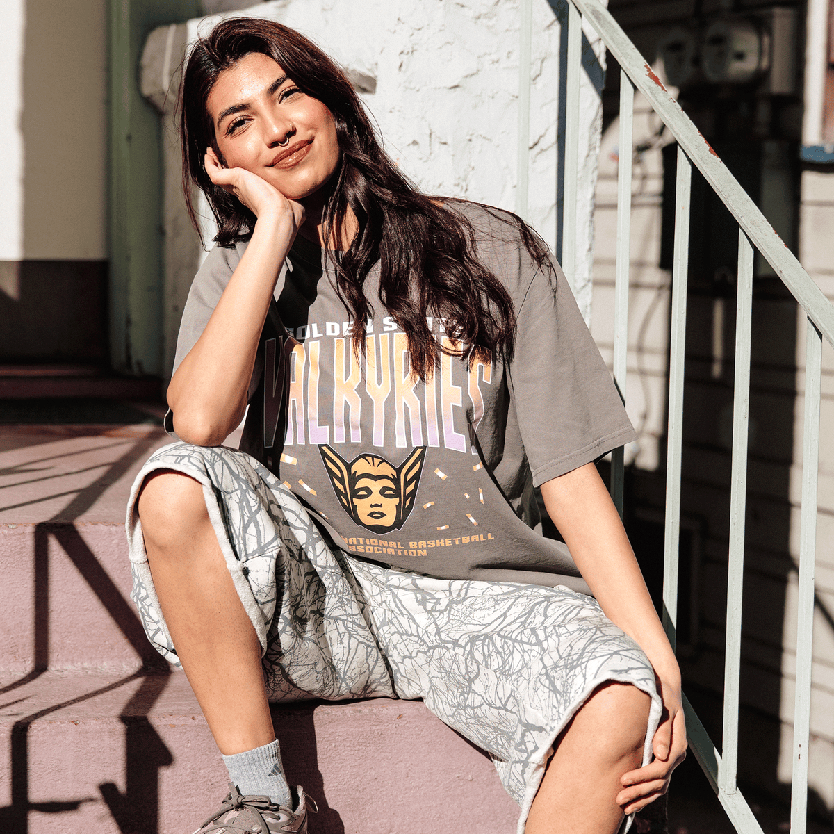 A woman with long dark hair and a septum piercing wears a GSV Women’s First Flight Tee and light-colored shorts with a dark, web-like pattern, sitting on outdoor concrete steps with her head leaned on her hand as sunlight casts deep shadows.