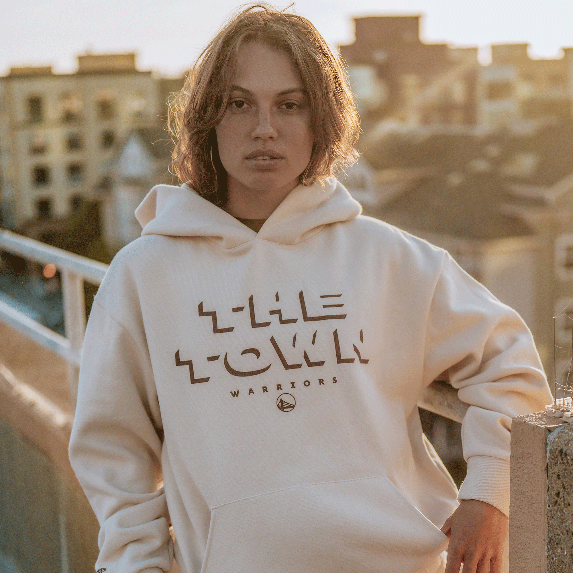 A person with short, wavy brown hair wears a loose white Ghost Block Letters Hoodie by GSW, leaning casually against a concrete rooftop railing at sunset with blurred city buildings in the background.
