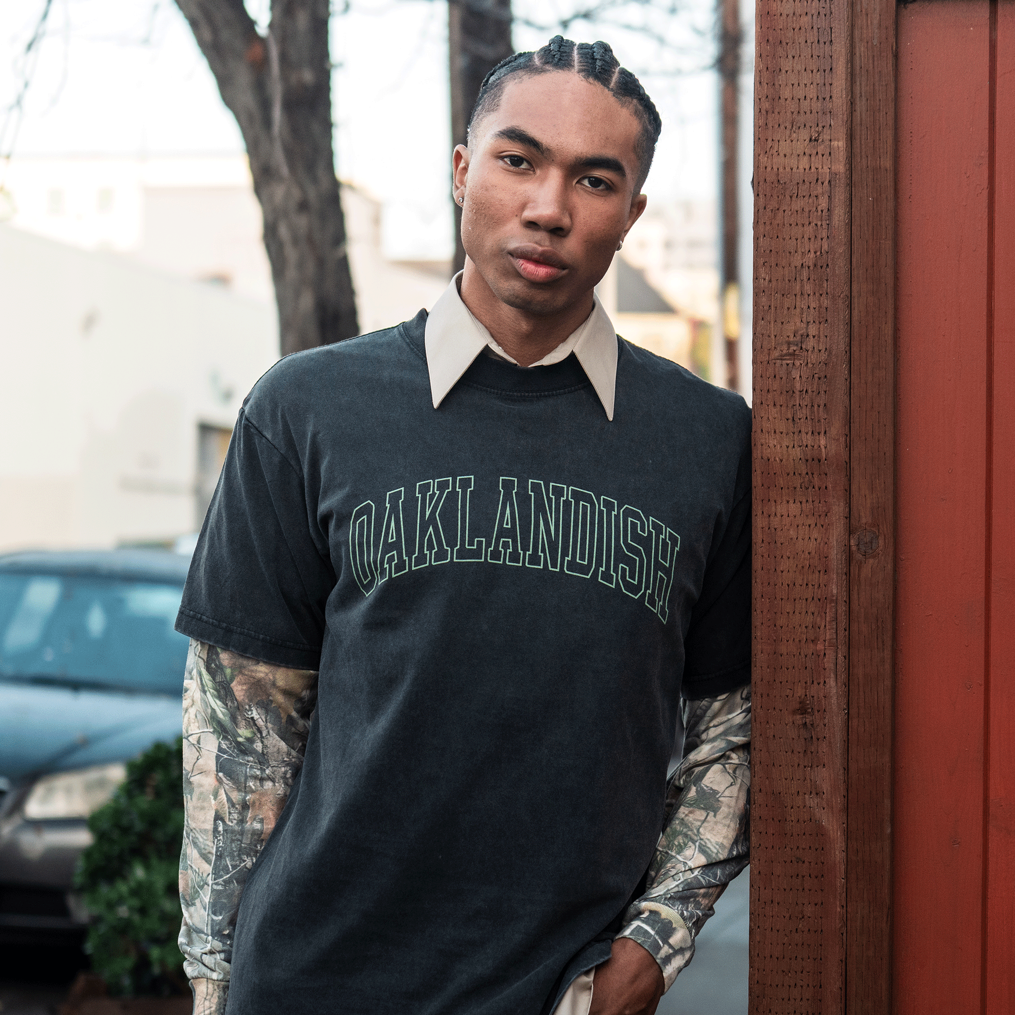 A young man with braided hair stands outside by a wooden fence, wearing the Oaklandish Hunter LS Tee layered over a beige collared shirt. A tree, car, and blurred buildings appear in the background.