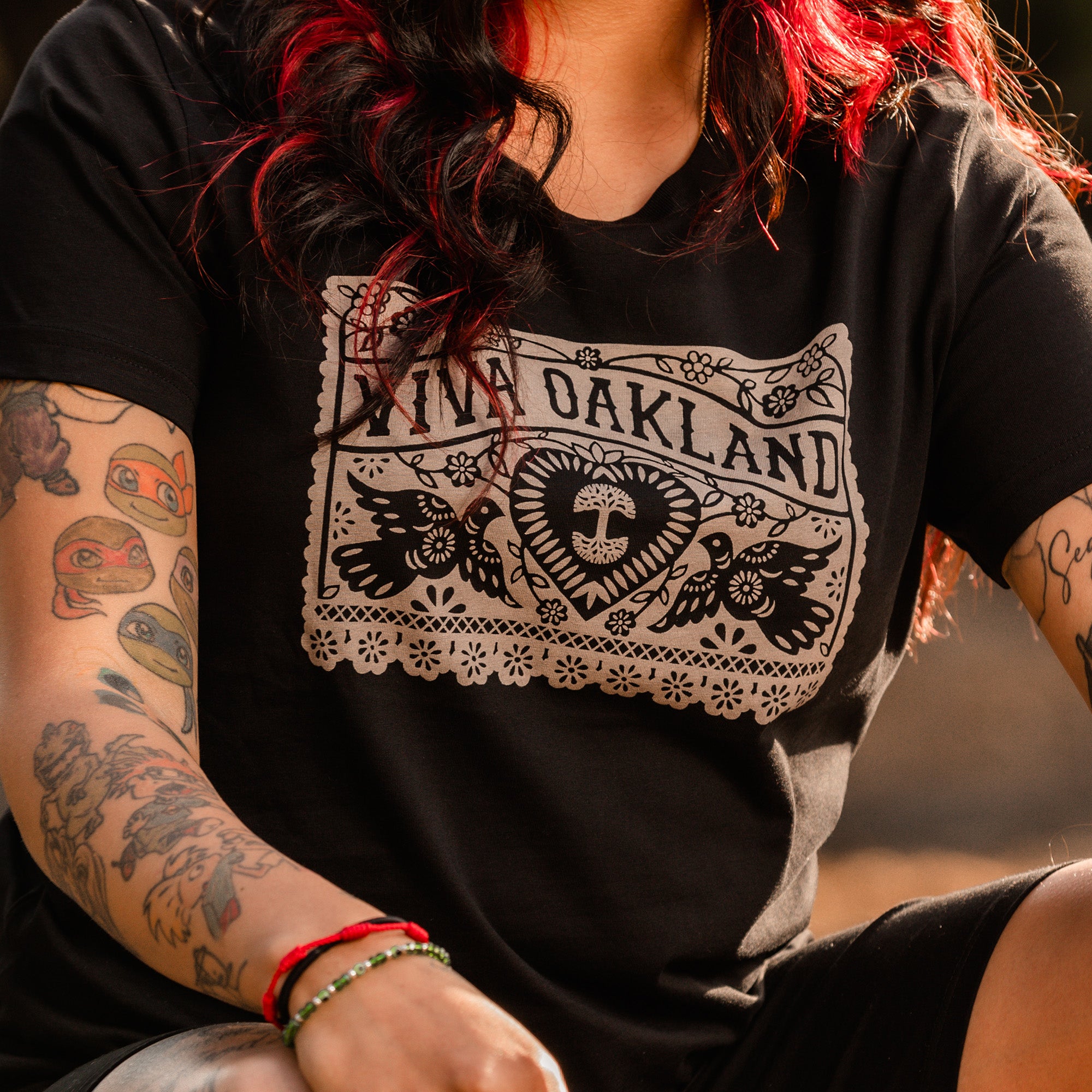 A person with long, wavy hair dyed black and red is wearing the Women's Viva Oakland Tee by Oaklandish. This classic fit t-shirt, featuring an intricate design with the text "Viva Oakland," includes a heart, flowers, and doves. Made of 100% cotton, the shirt beautifully complements their arms adorned with multiple colorful tattoos.