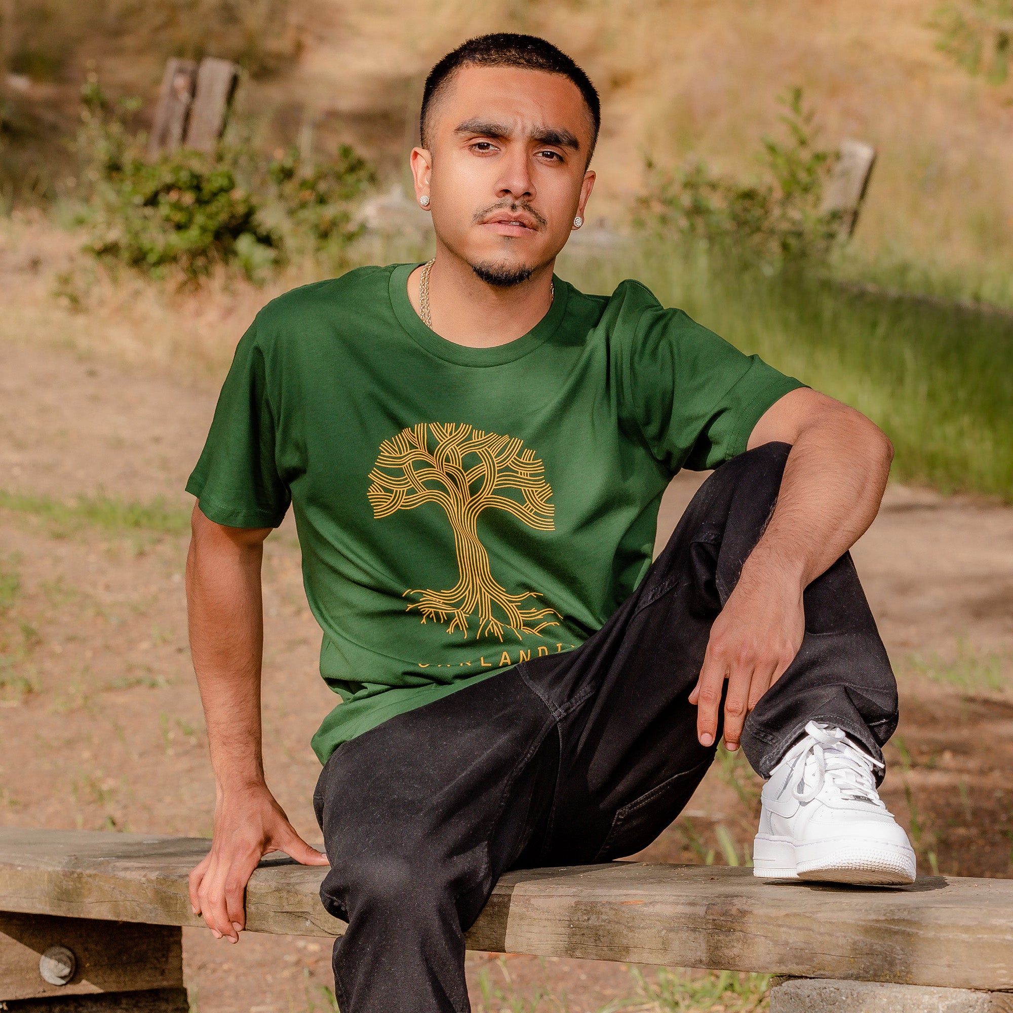 A man with short dark hair and a trimmed beard is sitting on an outdoor bench. He is wearing an Oaklandish Classic Logo Tee featuring a golden tree design, along with black pants and white sneakers. The background showcases a natural setting with grass, bushes, and wooden remnants.
