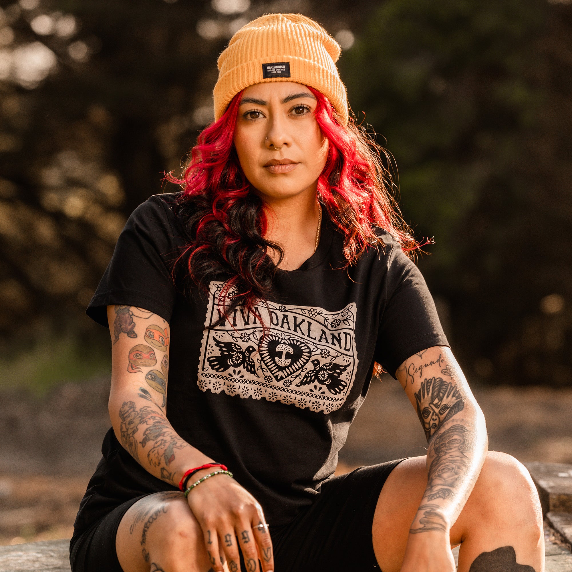 A person with bright red and black hair, dressed in a yellow beanie and an Oaklandish Women's Viva Oakland Tee made from 100% cotton with a classic fit, is sitting outdoors. Their arms and legs display prominent tattoos, including a spider web and various other designs. With a contemplative expression, they rest their hands on their knees.