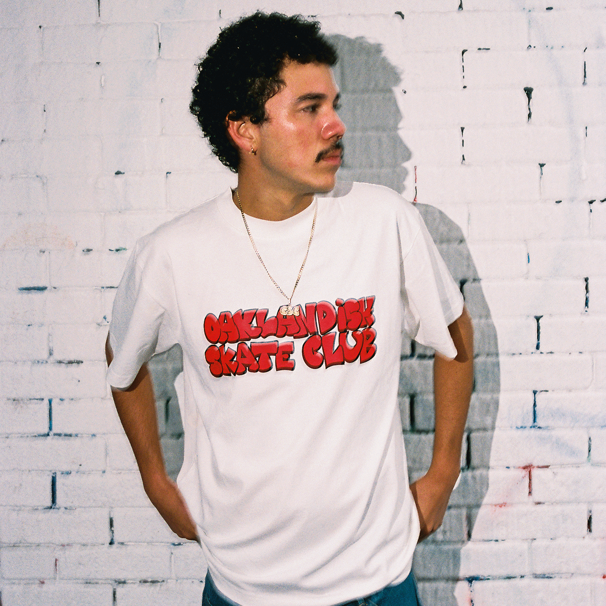 A person with curly hair and a mustache leans on a white brick wall, looking left. They wear the Oaklandish Skate Club Tee by Oaklandish, featuring red, cartoon-style text inspired by skateboard culture, hands behind their back for a casual look.