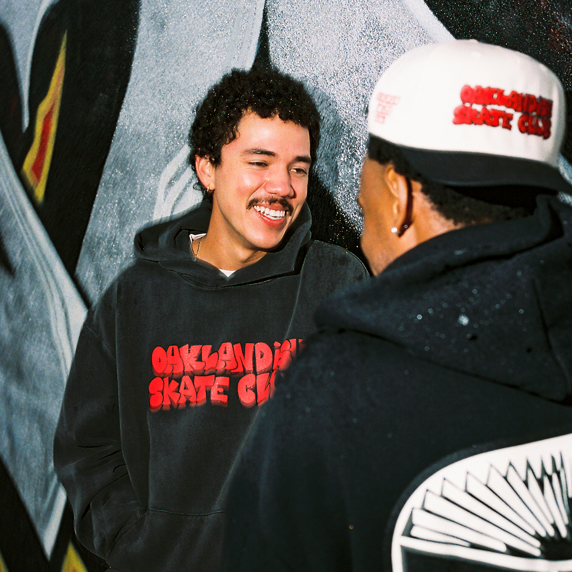Two people smile and chat in front of a graffiti mural, both wearing Oaklandish Skate Club Hoodies from Oaklandish, featuring “OAKLANDISH SKATE CLUB” in red text—a joyful nod to urban street style.