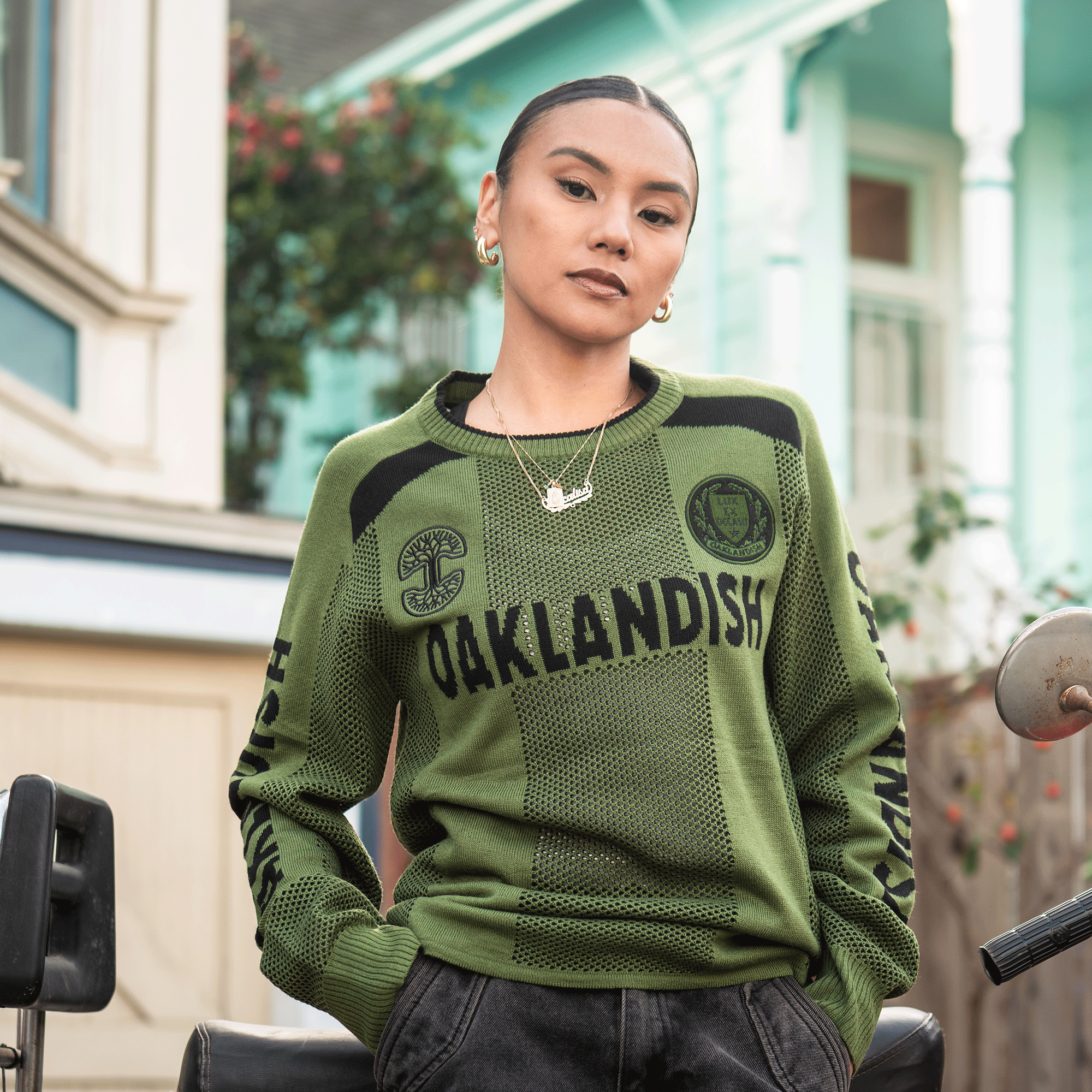 A woman with slicked-back hair and gold jewelry stands confidently before pastel houses, wearing the Oaklandish Terrace Sweater—a green, unisex knit by Oaklandish. She looks calmly at the camera, hands in her pockets.