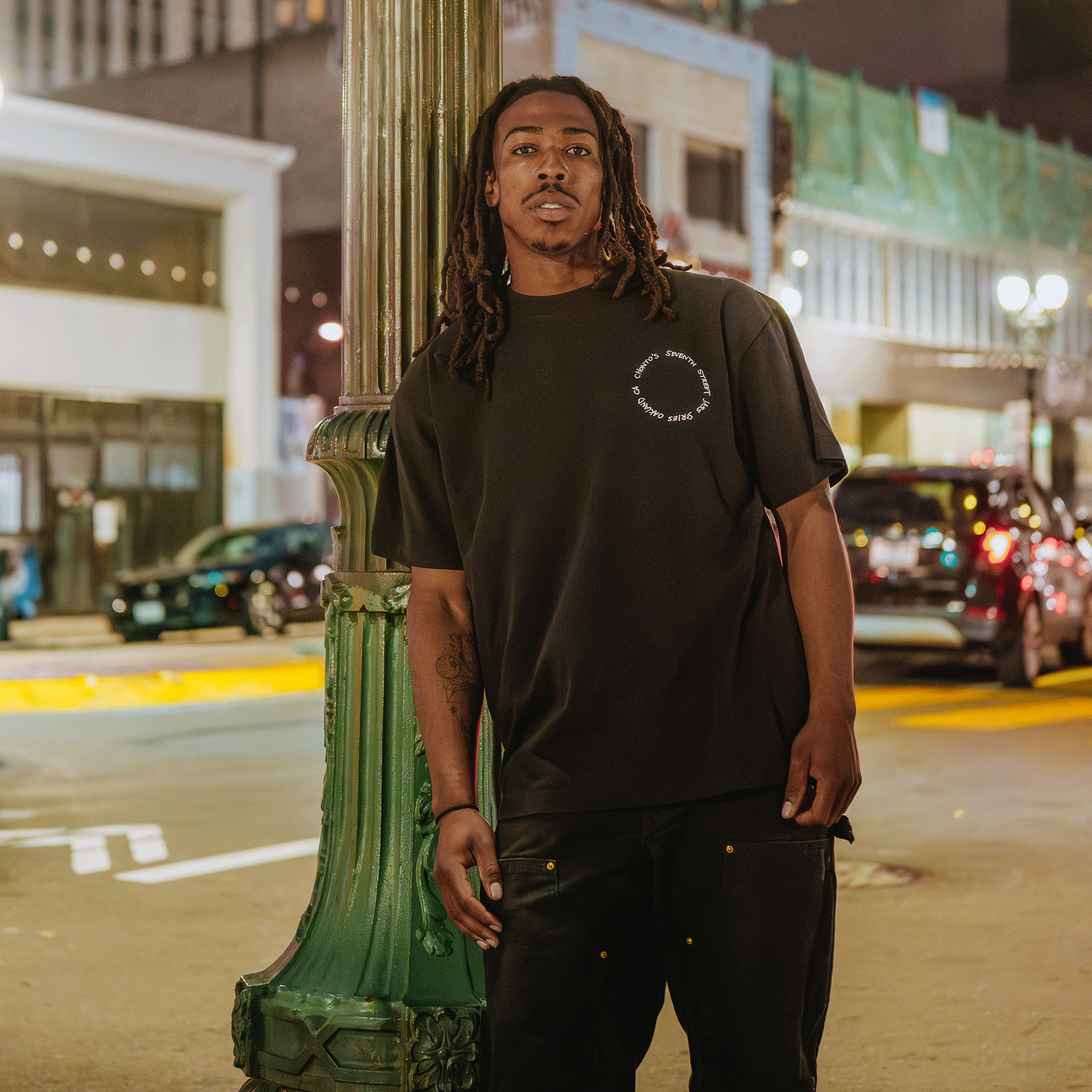 Man standing on a city street at night, wearing a black t-shirt and pants.
