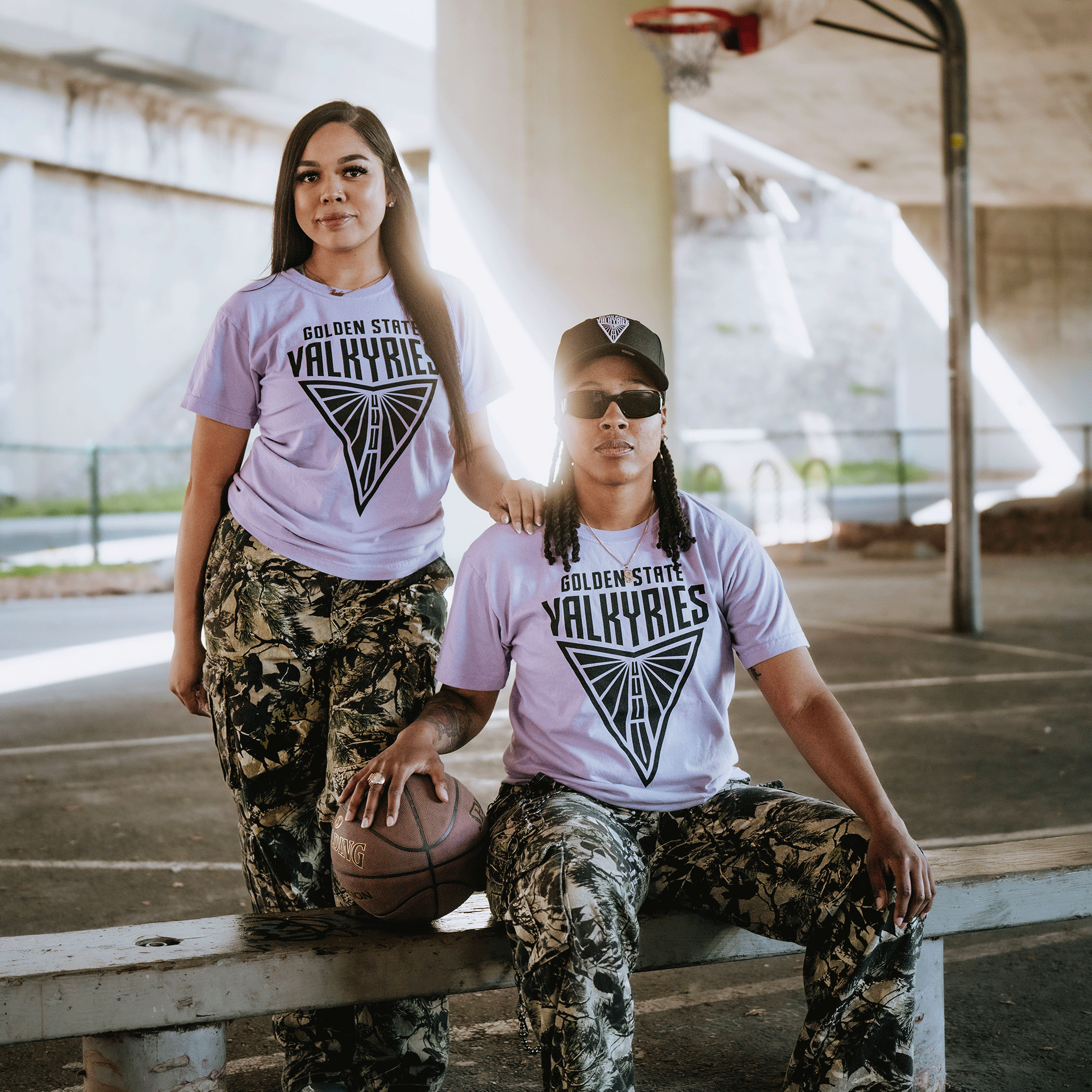 Two people pose under a bridge in matching GSV Golden State Valkyries Primary Premium Tees and camo pants. One stands with hands on hips; the other, in sunglasses and cap, sits on a bench with a basketball.