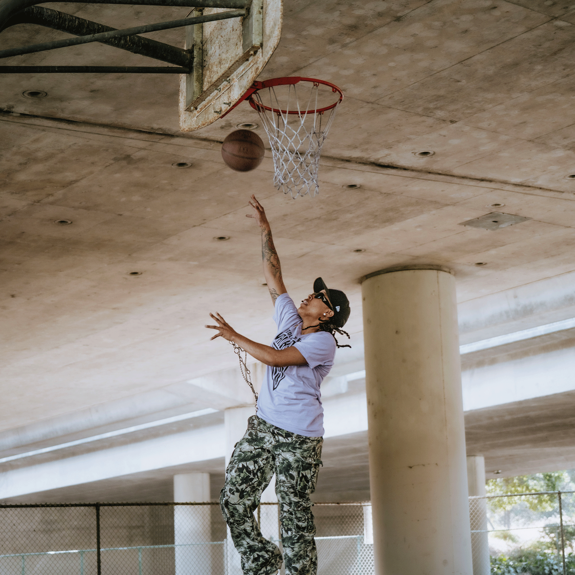Wearing a GSV Golden State Valkyries Primary Premium Tee, camouflage pants, and a black cap, a person leaps for a basketball layup under an overpass, arm outstretched as the ball nears the hoop amid urban concrete pillars.