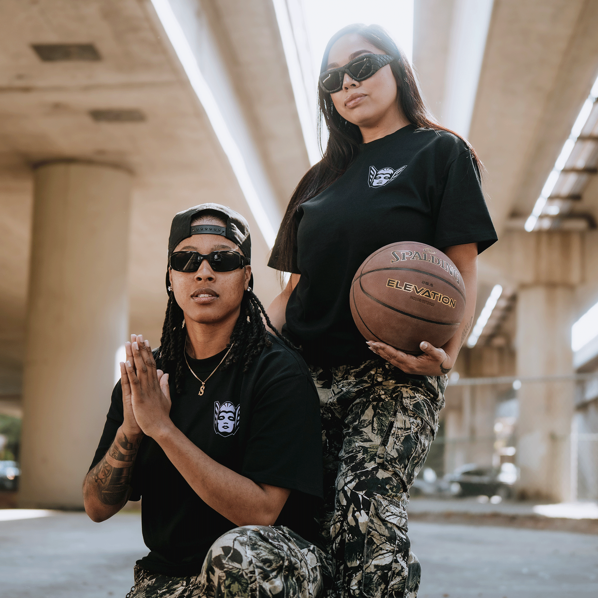 Two people pose under a concrete overpass in GSV Golden State Valkyries Renowned Heavy Tees and camo pants. One kneels, the other stands holding a basketball, both wearing dark sunglasses as sunlight streams down above.